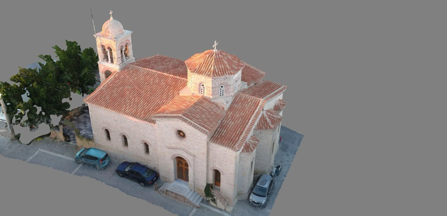 A church with stone walls and terracotta roof tiles, a small bell tower on the left, and a central dome with a cross. There are parked cars on the street in front, and trees on the left side.