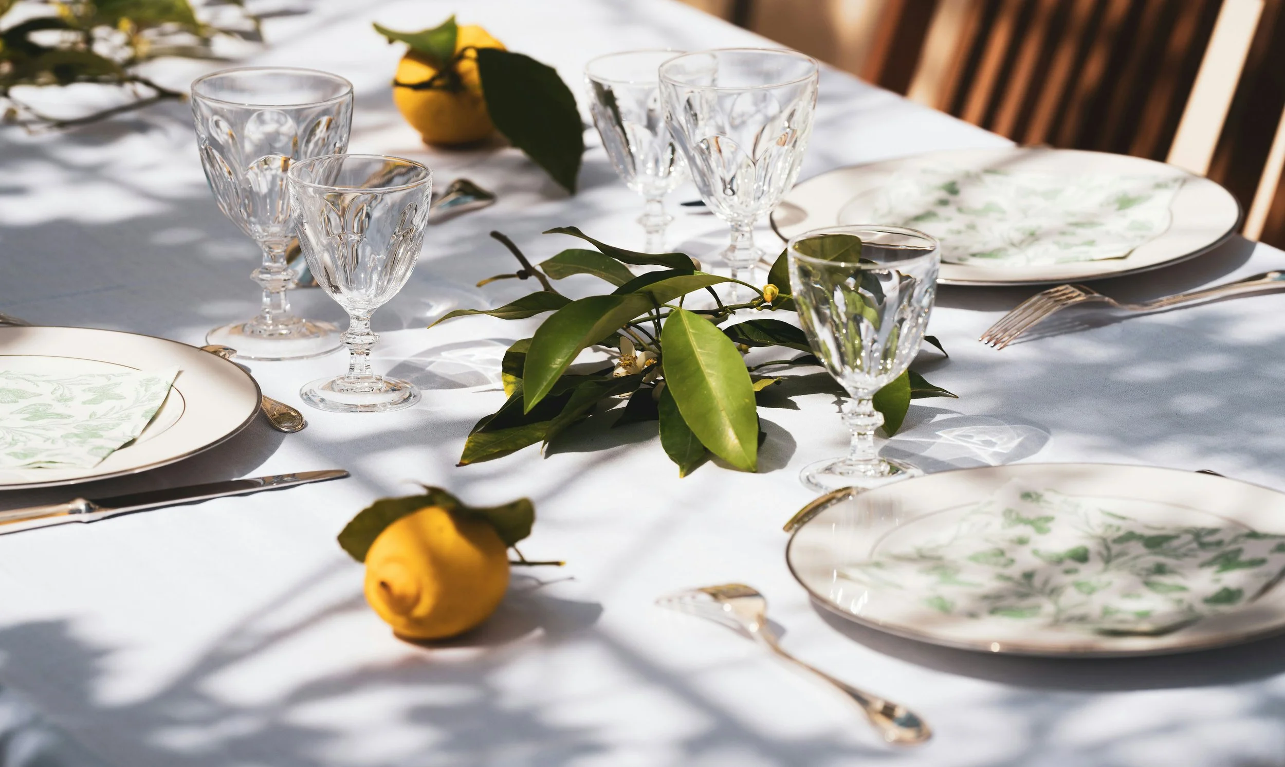 Elegant table setting with white tablecloth, floral-patterned plates, crystal glasses, silverware, greenery, and yellow lemons under sunlight.