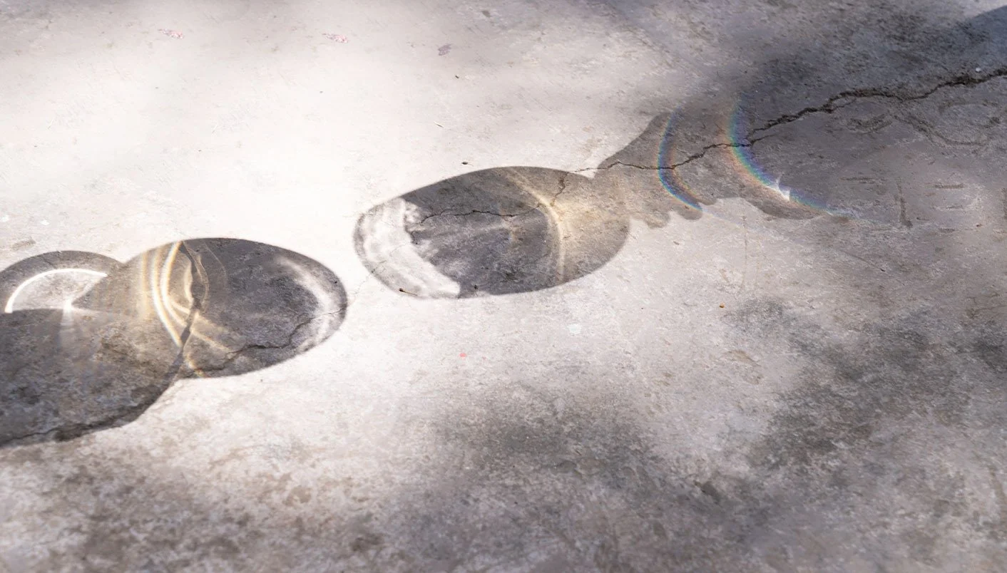 Shadows of three round objects cast on a cracked concrete surface, with a rainbow-like light distortion.
