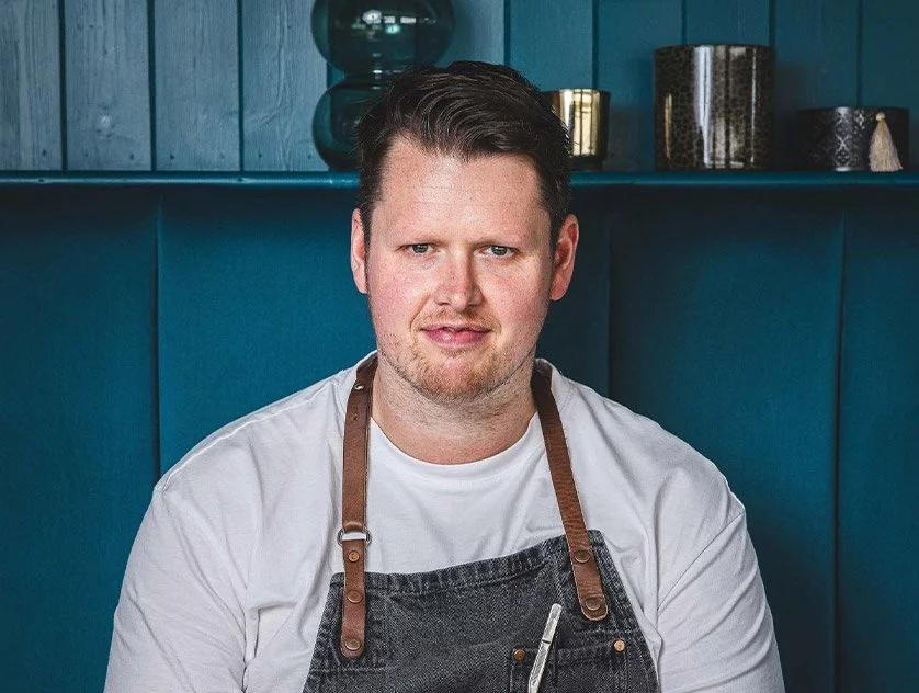 A man in a white t-shirt and apron sitting in front of a blue wooden wall with decorative objects on shelves.