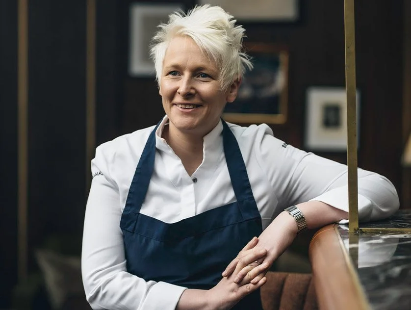 Woman with short platinum blonde hair wearing a white chef's coat and navy apron, smiling and leaning on a kitchen counter.