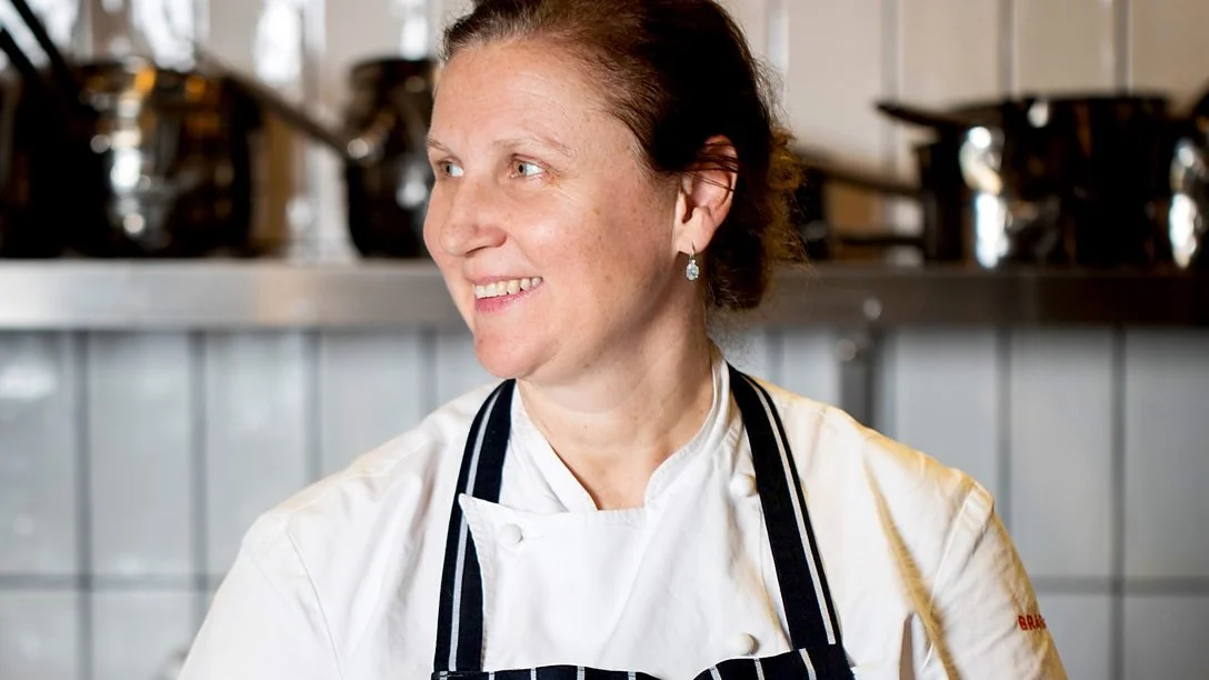 A woman chef in a white uniform and black apron smiling in a kitchen with stainless steel equipment.