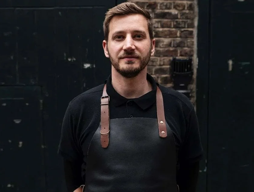 Man with short brown hair and beard wearing a black shirt and leather apron standing outdoors in front of a black door and brick wall.
