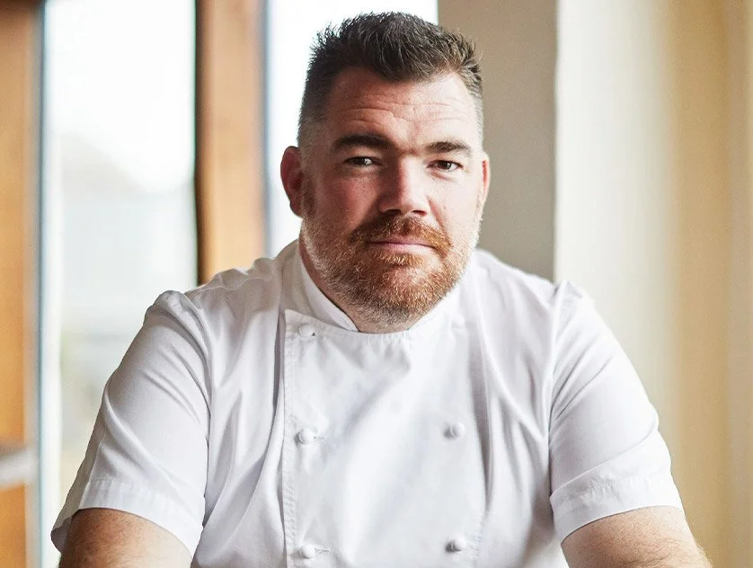 A male chef with short dark hair and a beard, wearing a white chef's jacket, sitting indoors near a window with a neutral expression.