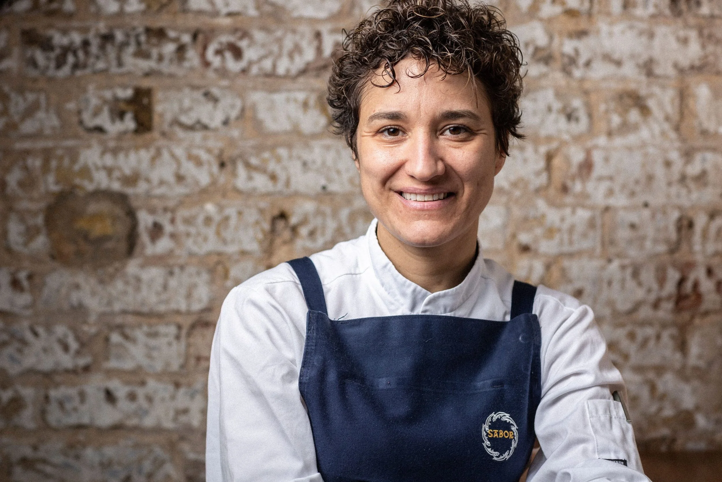 A female chef smiling, wearing a white chef coat and a navy apron with a logo that says "Sabor" on it, standing in front of a brick wall.