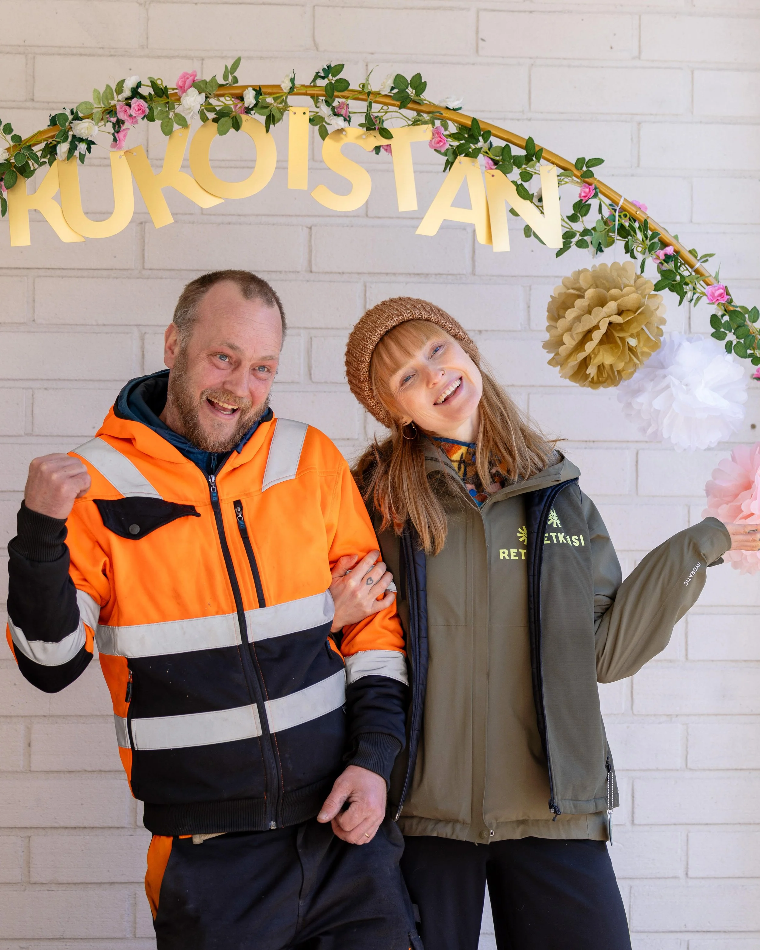 Kaksi nauravaa kävijää poseeraa kukin ja paperipomponein koristellun kultaisen Kukoistan-kaaren alla Kukoistus-tapahtumassa.