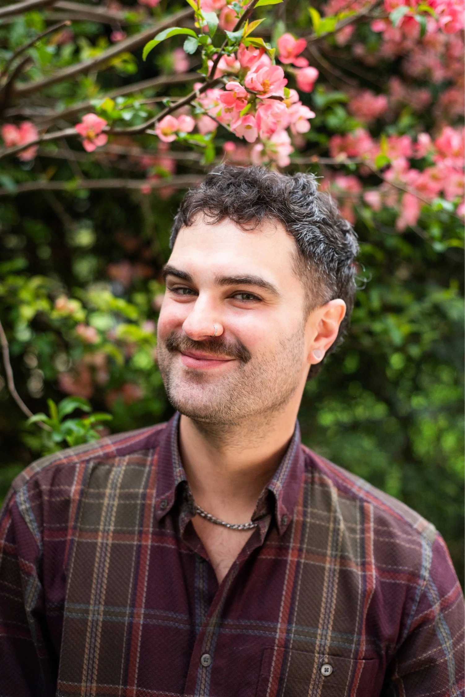 Man with brown short hair smiling. Nose ring, mustache and stubble. Flannel shirt on, with plants and flowers in the background blurred.