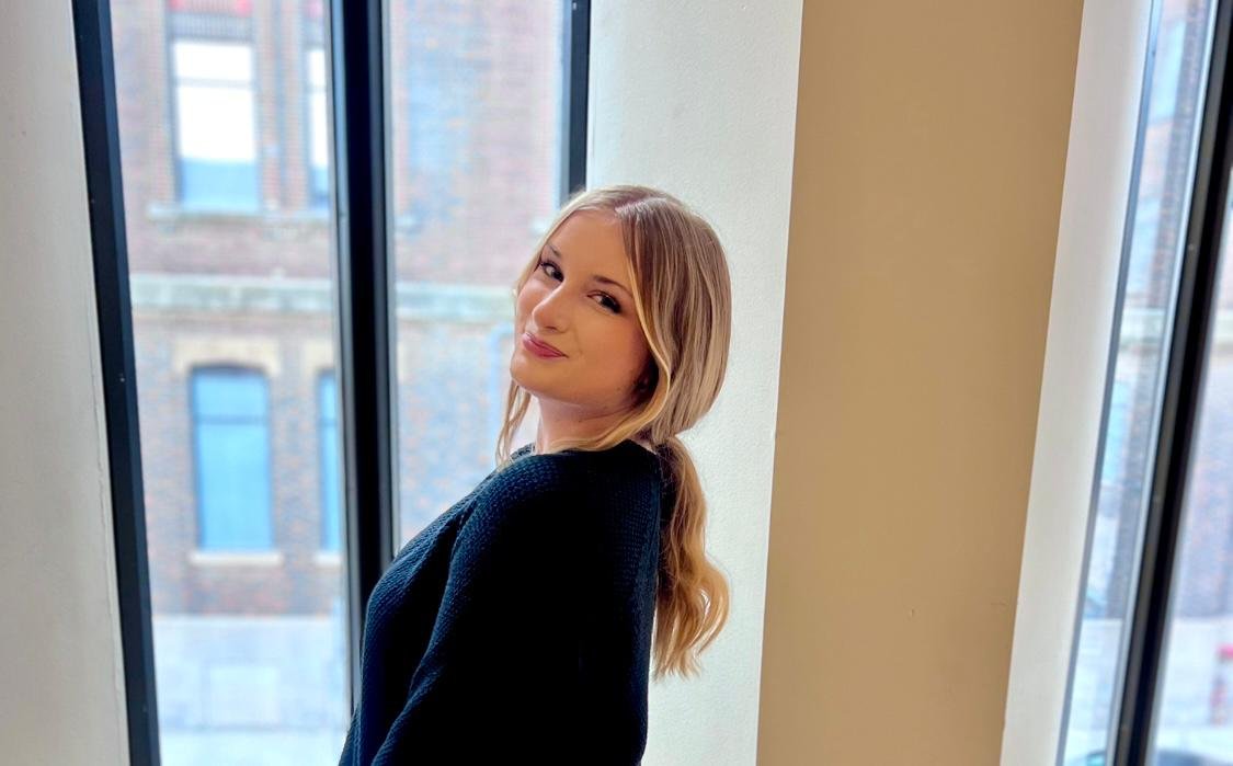 Young woman with blonde long hair and a blue sweater smiling with a window behind her