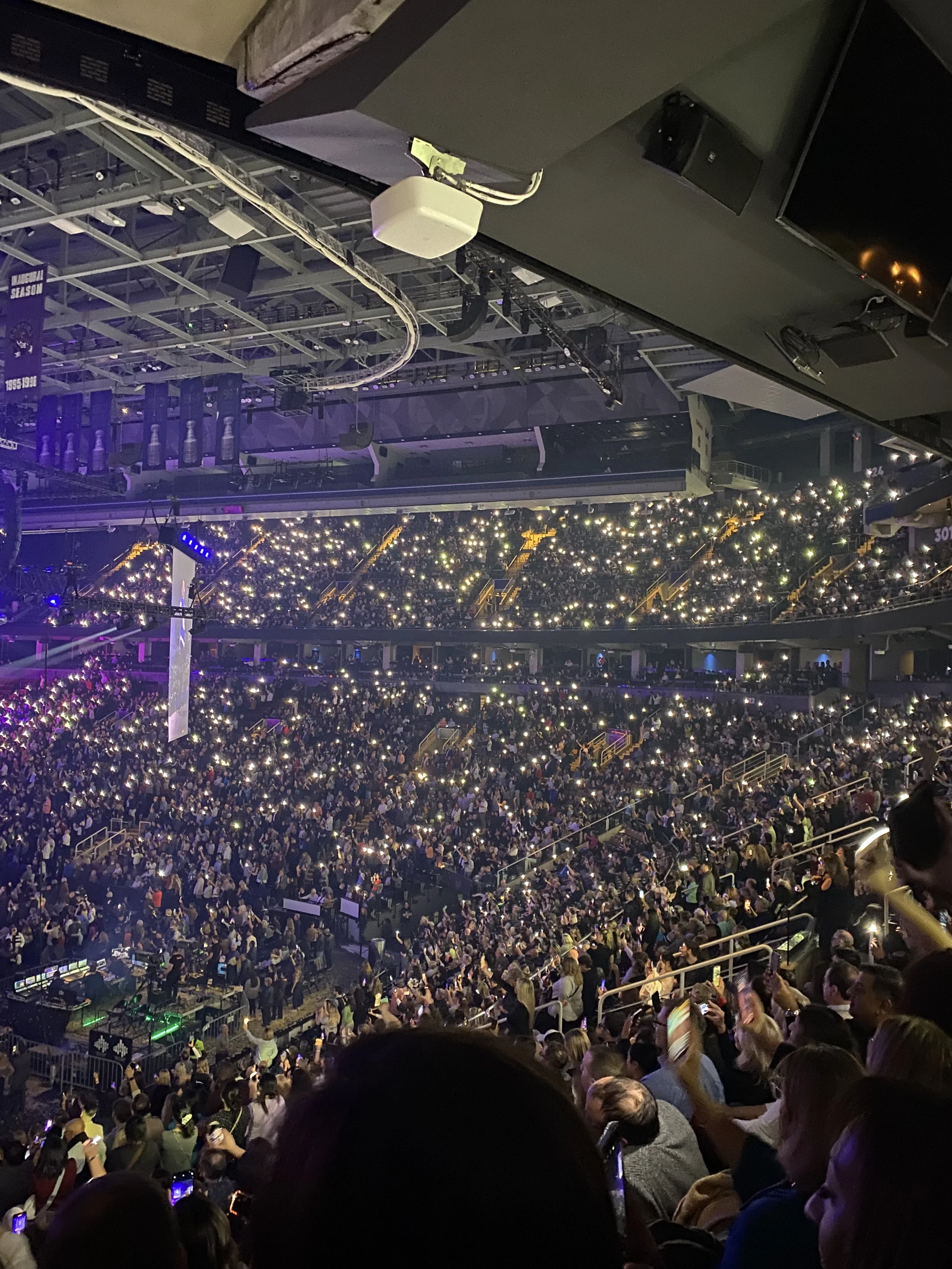 A large indoor concert venue filled with a crowd of people, many holding up phones with flashlights on, and stage lighting effects suspend over the audience.
