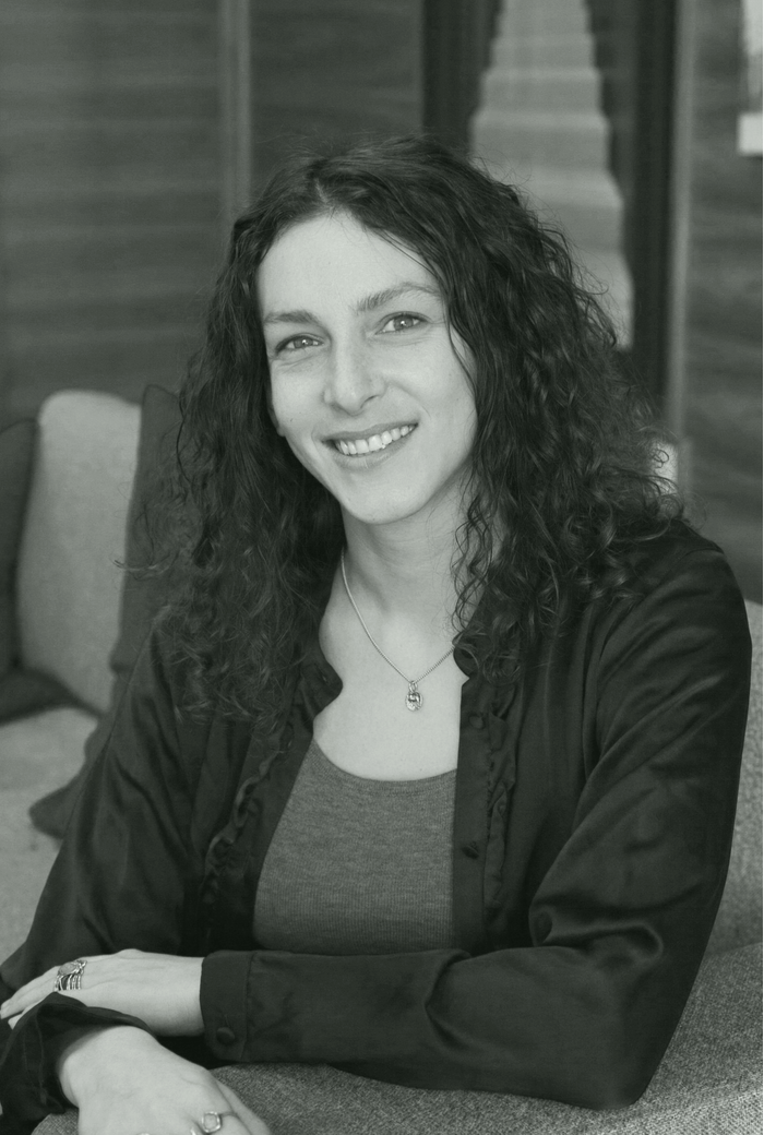 A woman with curly dark hair smiling at the camera, sitting at a table in an indoor setting with wooden paneling in the background.