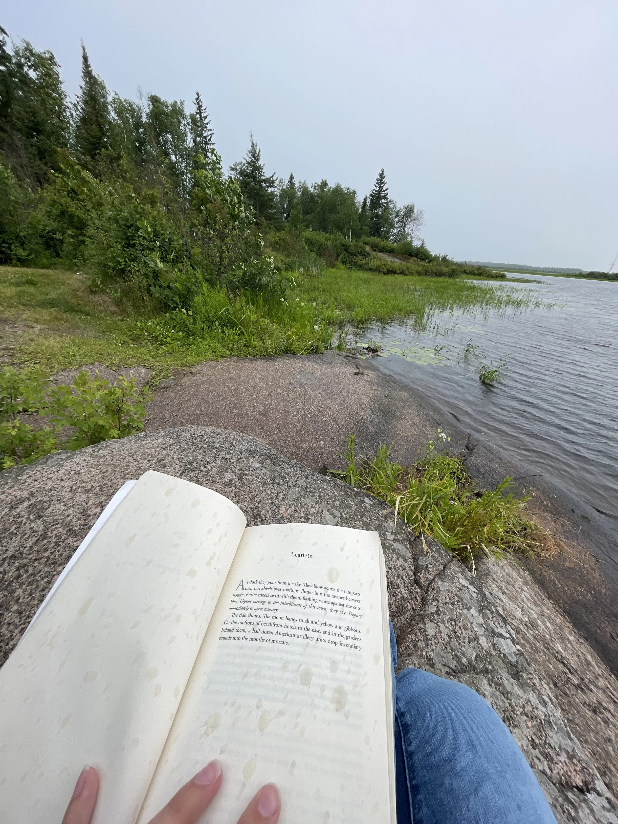 Person sitting on rocks by a river, reading a book with trees and water in the background.