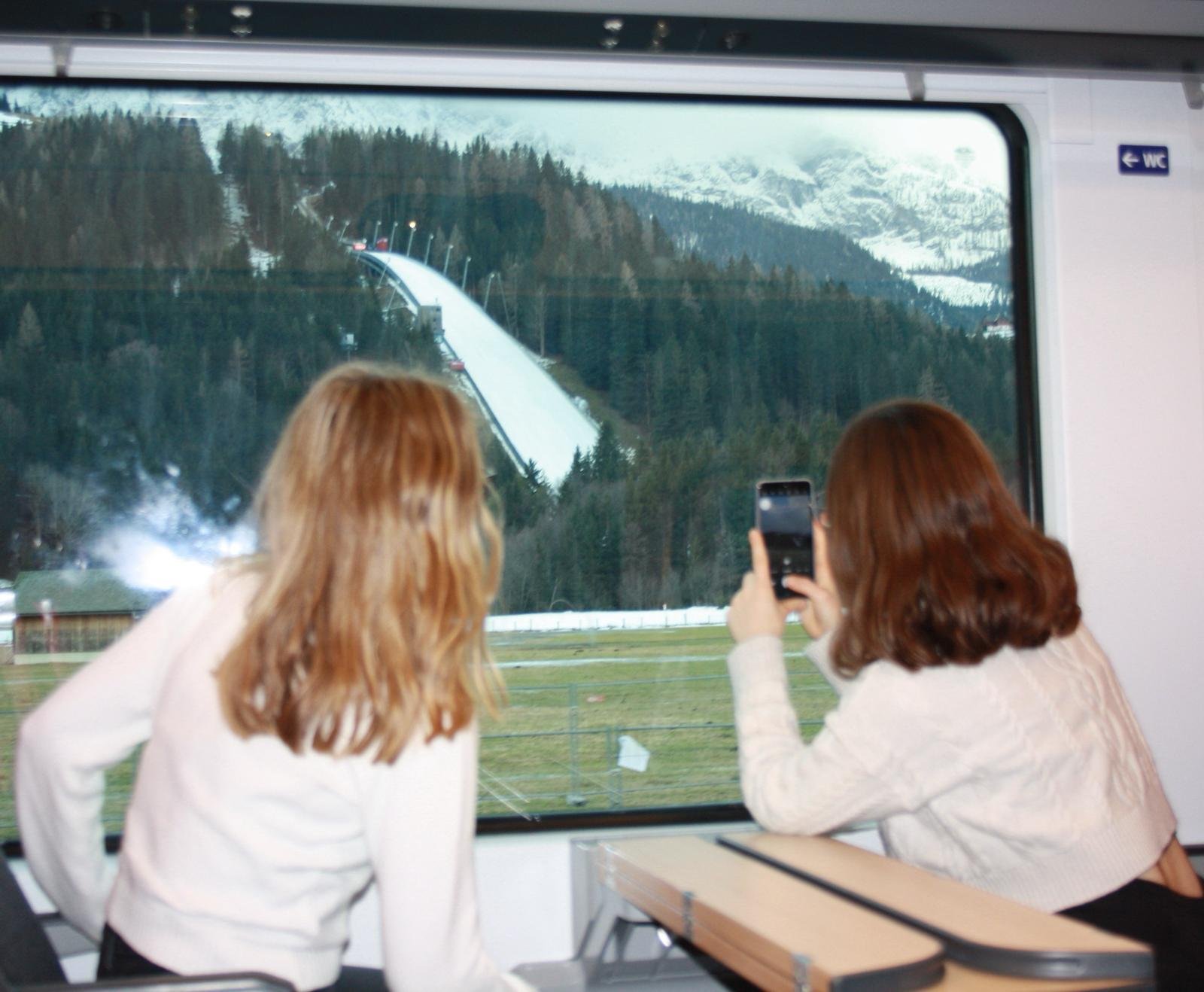 Two women with red hair looking out a train window at a snowy mountain landscape with a ski jump on a forested hillside.