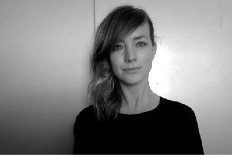 A woman with shoulder-length, wavy hair standing against a plain wall, wearing a dark top, in black and white.