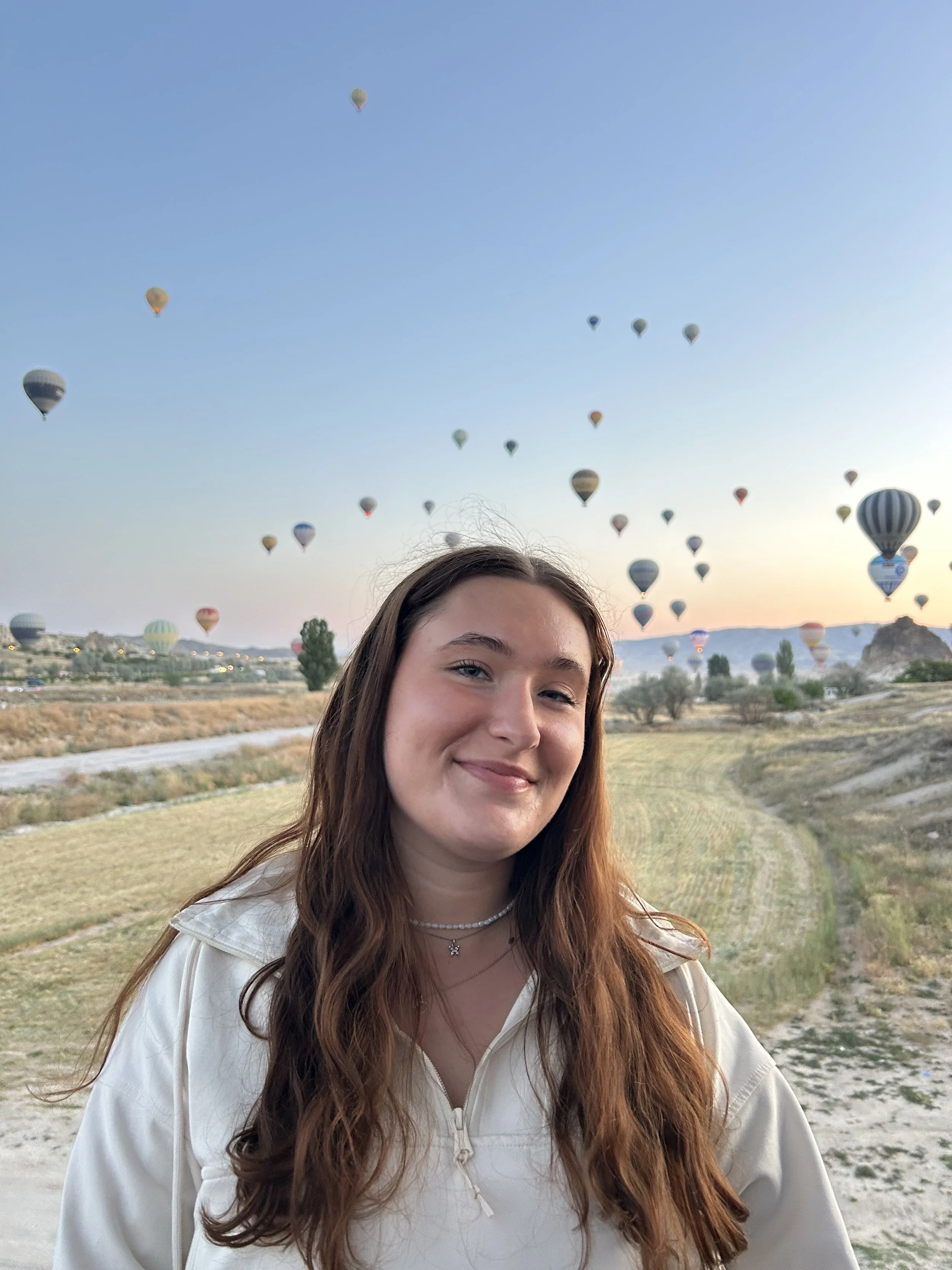 Young woman with long hair smiling outdoors with hot air balloons behind her