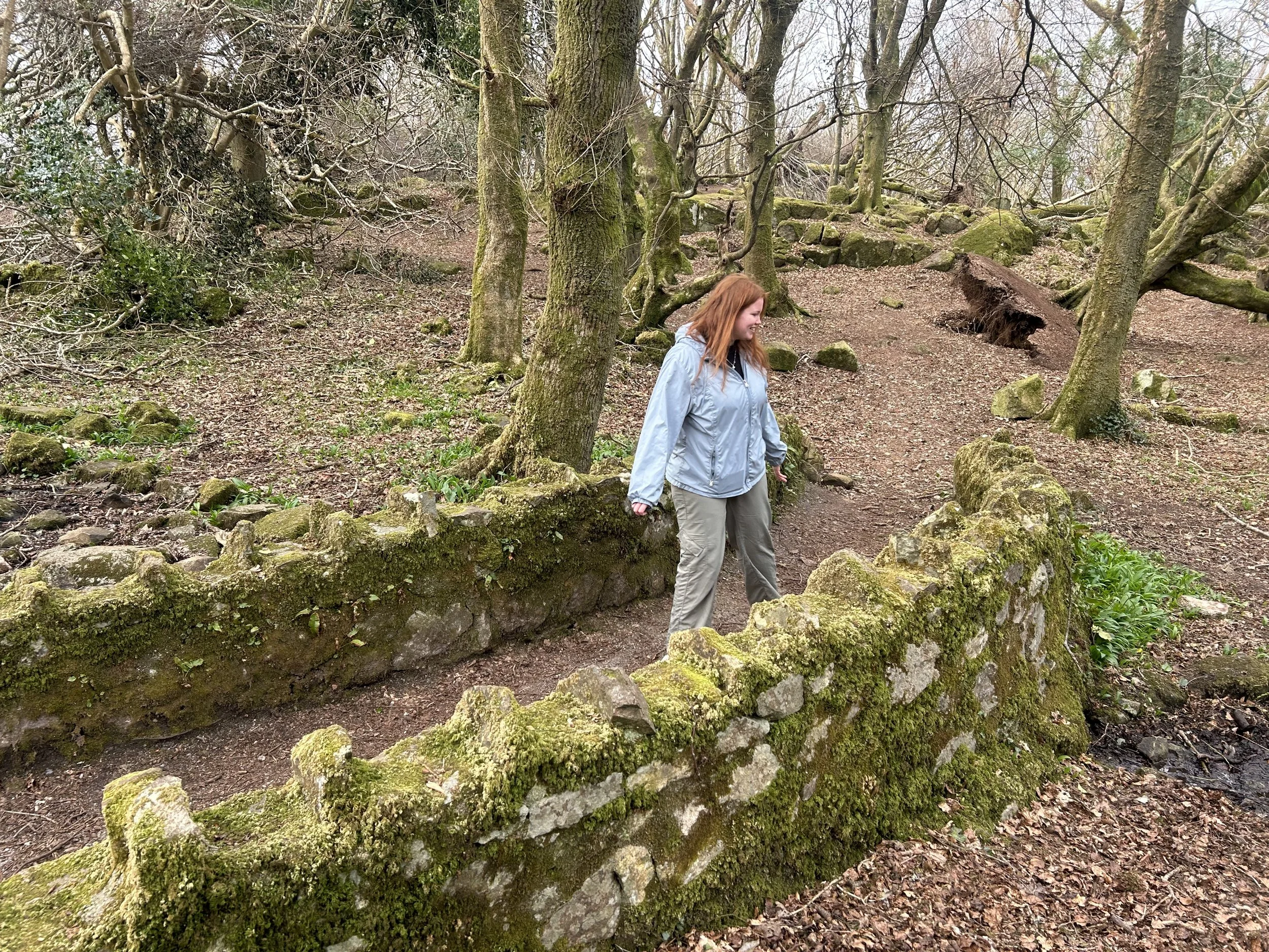 A woman with long red hair in a gray jacket and beige pants standing on a moss-covered stone wall in a wooded area. Trees and a dirt path are visible in the background.