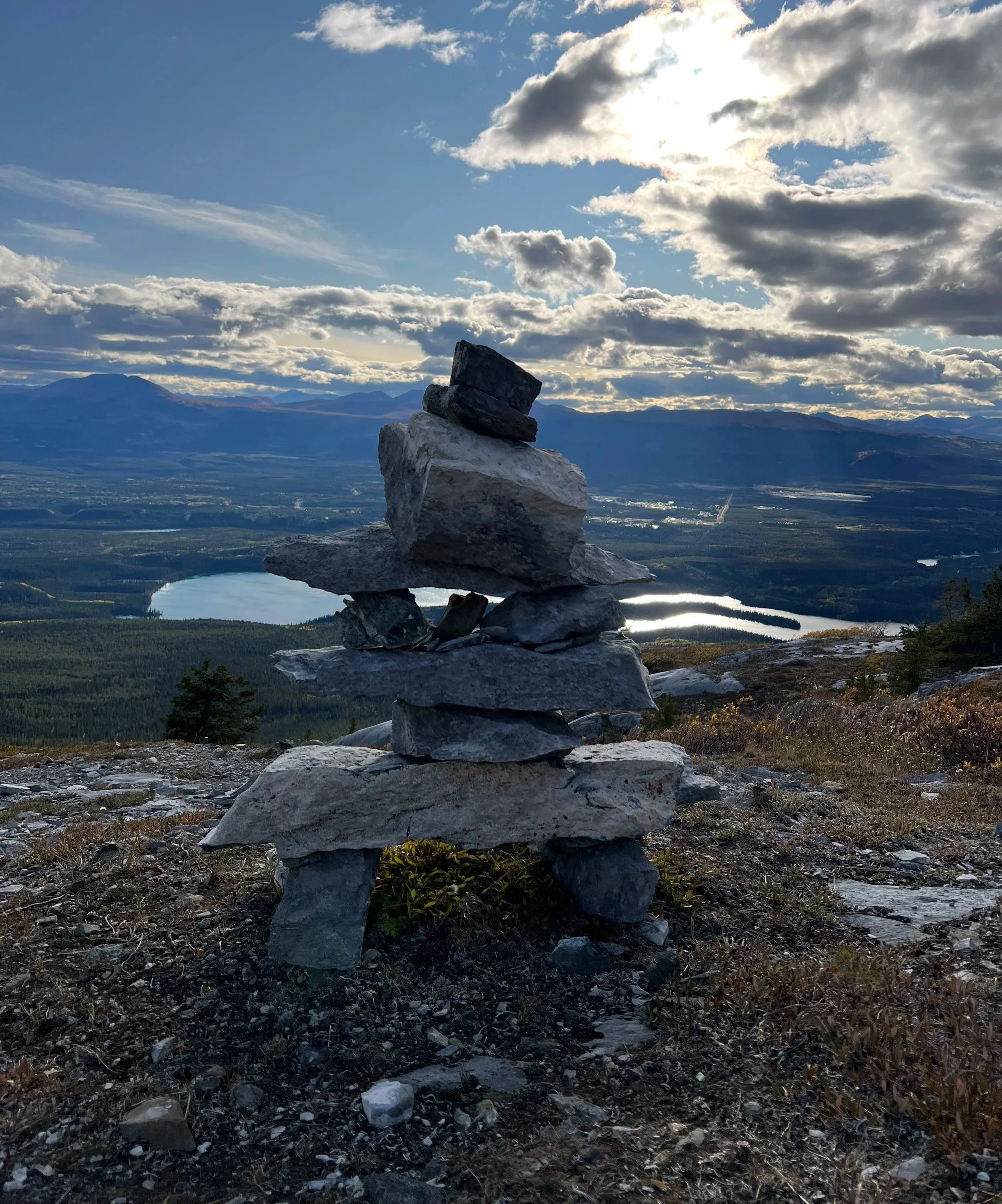Stacked stones on a mountain overlooking a lake and distant mountains under a cloudy sky.