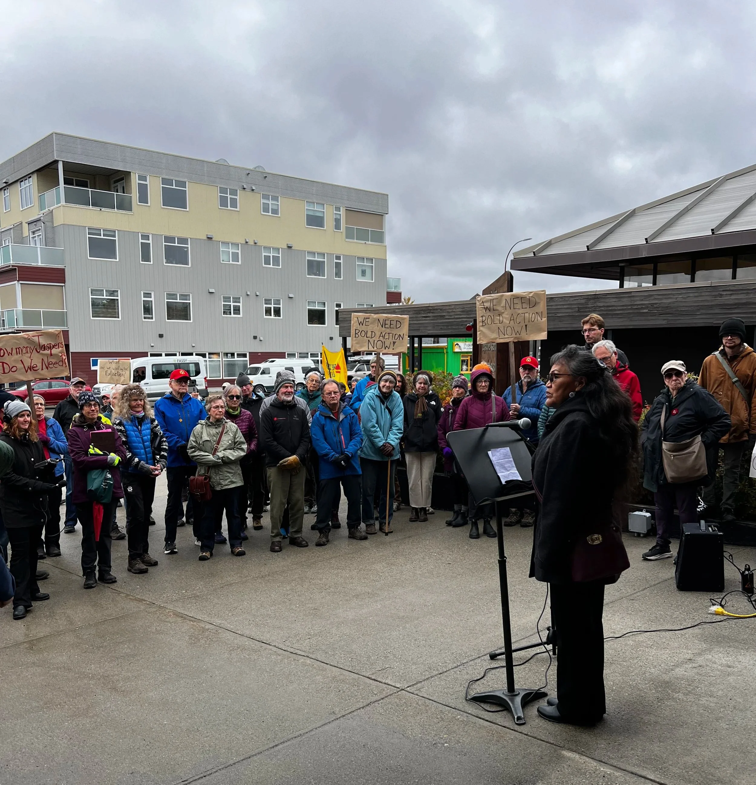 A woman speaking at a microphone during a protest, with a crowd of people holding signs behind her. The signs read "We need bold action now!" and other messages. The protest is taking place outside in front of modern apartment buildings on a cloudy day.