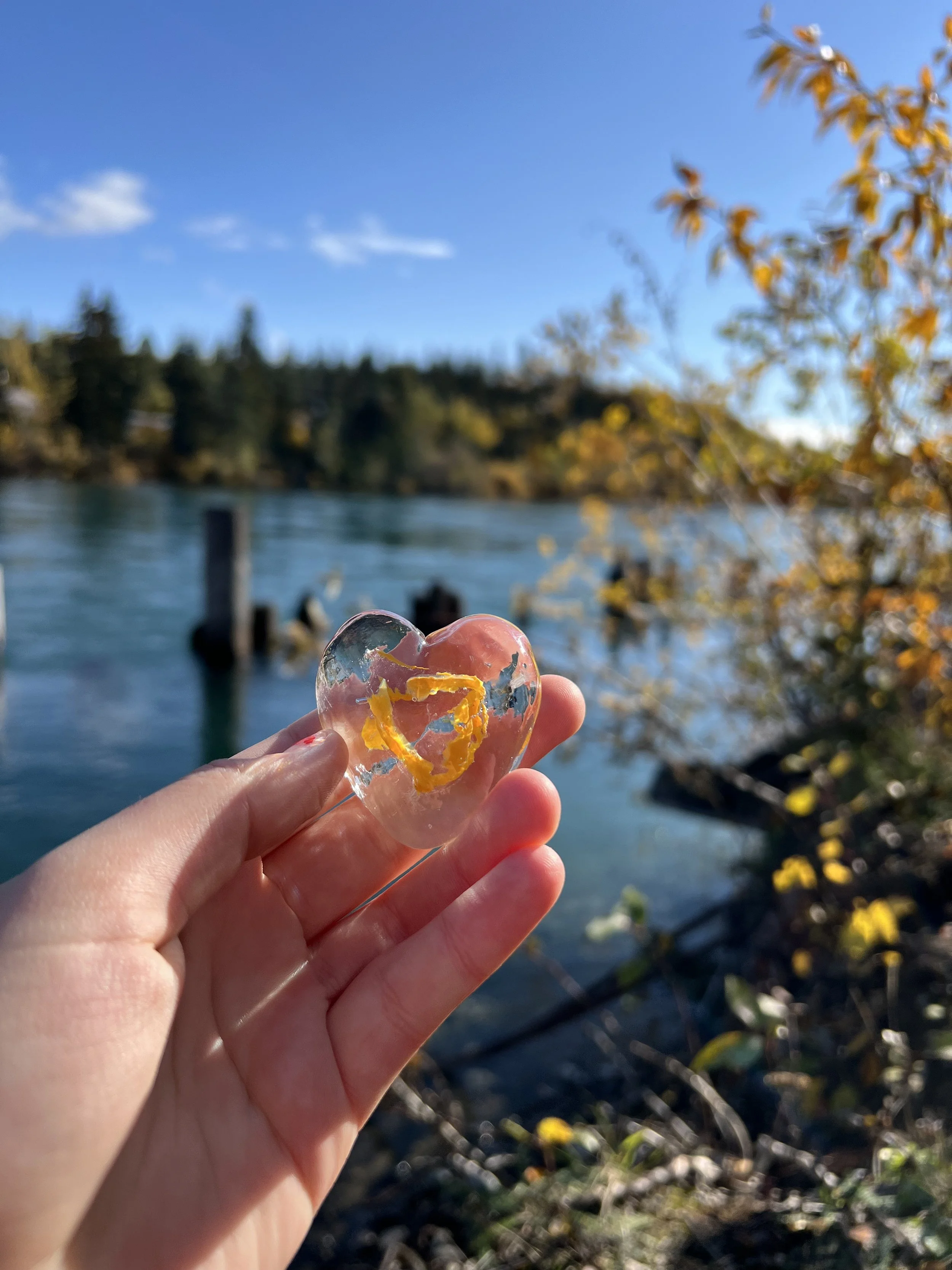 Hand holding a small, heart-shaped piece of clear glass with yellow and blue markings against a lakeside background with trees and a blue sky.