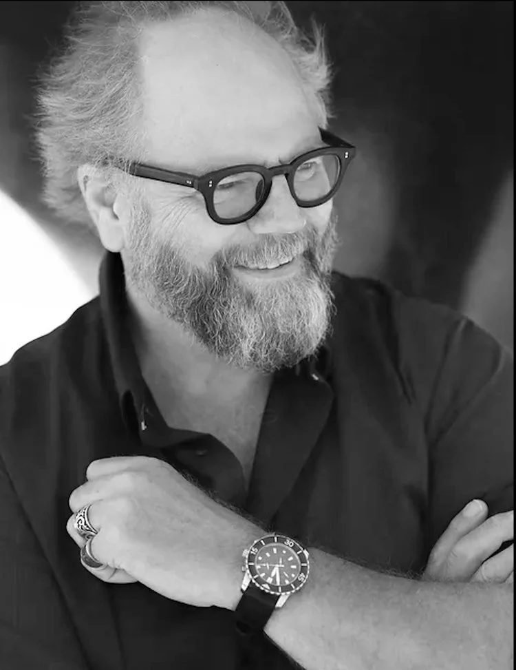 A smiling bearded man with glasses, wearing a dark shirt, with arms crossed, seated indoors.