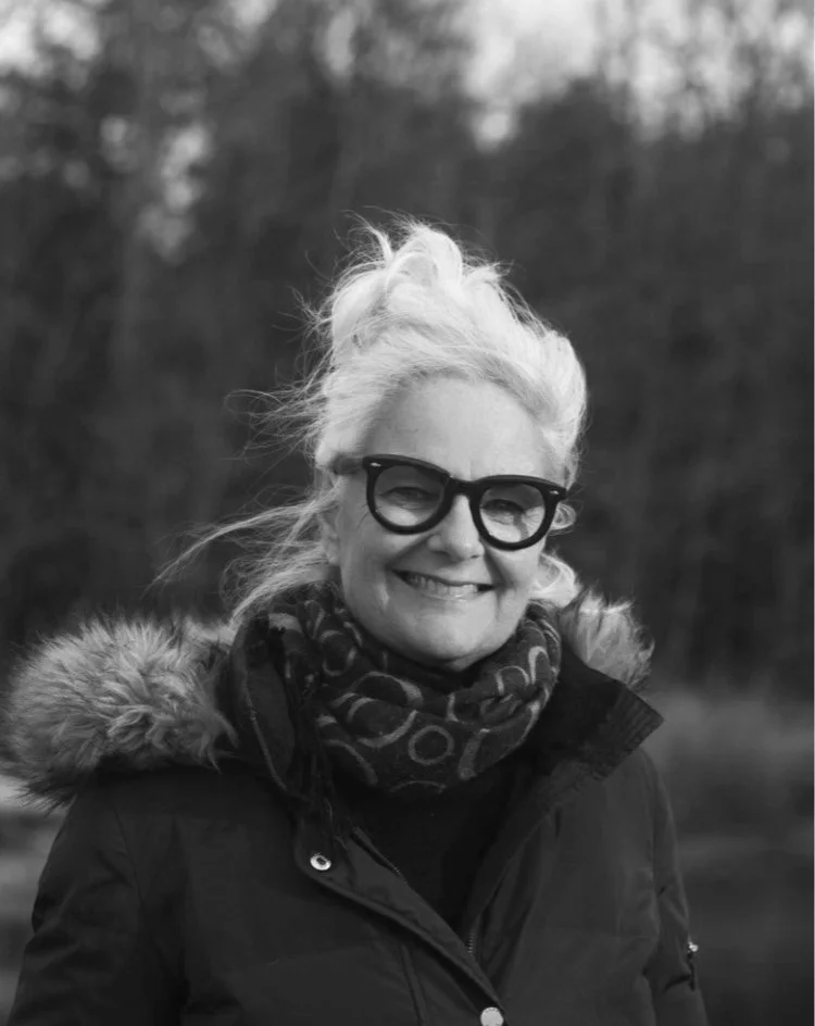 A black-and-white photo of a smiling woman with glasses and light-colored hair styled in a bun, wearing a winter coat with a fur-lined hood and a patterned scarf, outdoors with blurred trees in the background.
