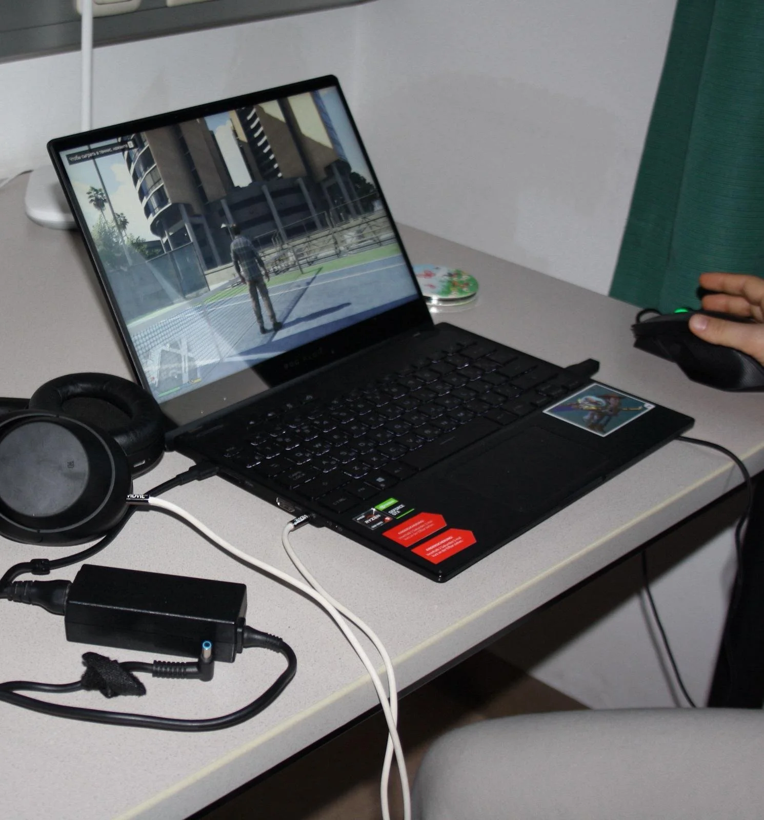 Laptop on a desk displaying a video game scene with a character standing outdoors near a building, with headphones and a hand using a computer mouse nearby.
