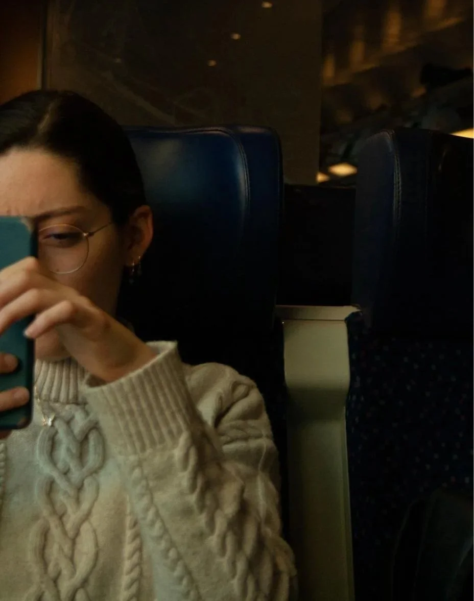 A person with dark hair and glasses taking a photo with a smartphone on a dark train or airplane, wearing a beige sweater with a cable knit pattern.