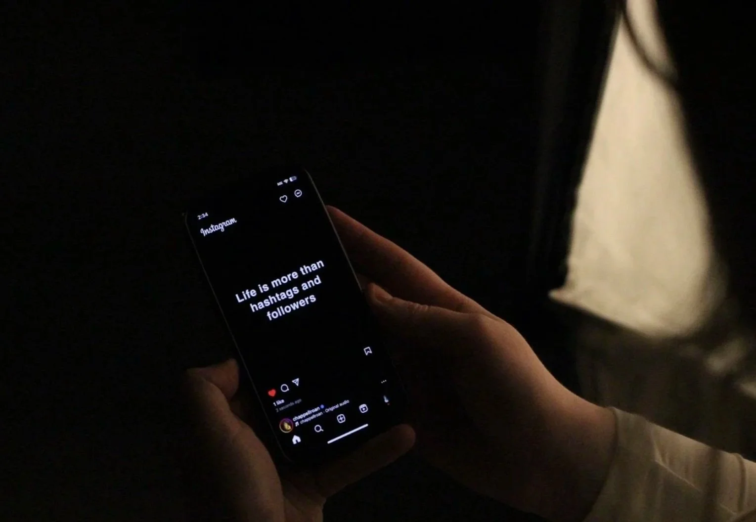 A person holding a smartphone displaying an Instagram post that reads, 'Life is more than hashtags and followers,' in a dark environment.