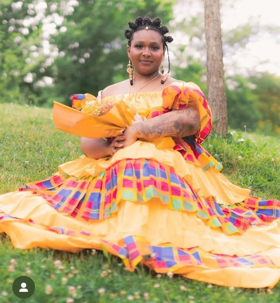 A woman sitting outdoors on grass, wearing a colorful, patterned dress with puffed sleeves, holding a bouquet of flowers, with trees in the background.