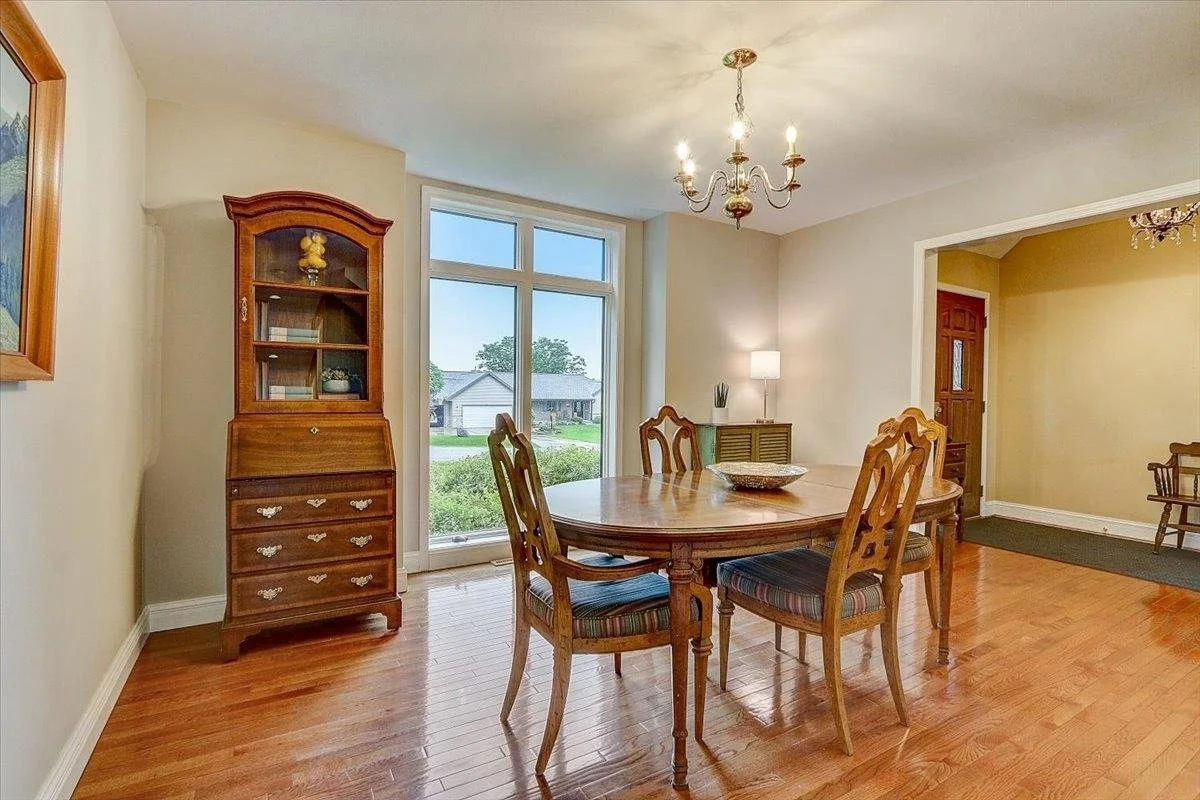 Dining room with hardwood floor, a wooden dining table with five chairs, a china cabinet with glass doors, a large window showing a suburban neighborhood, and a chandelier hanging from the ceiling.