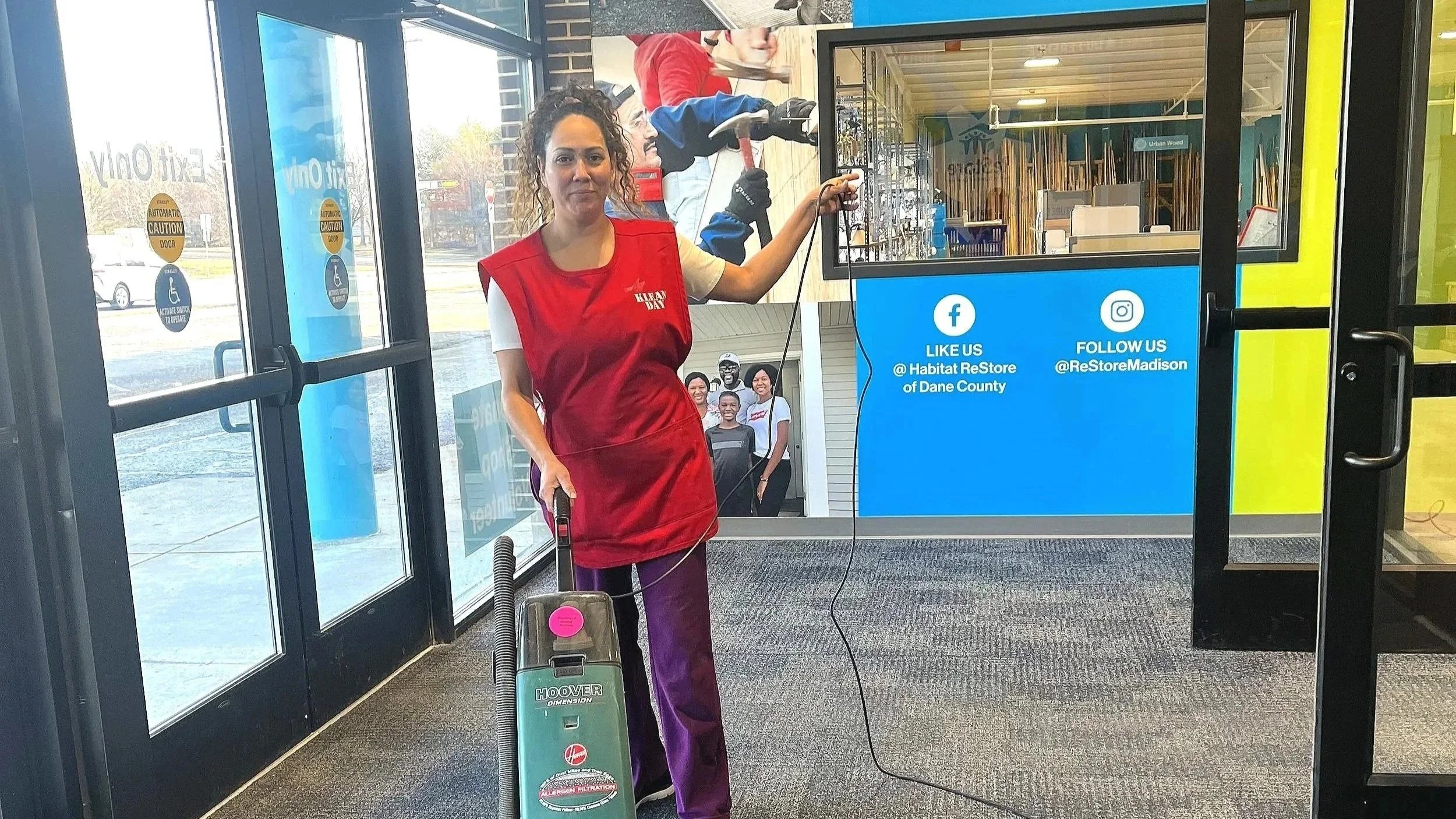 Woman in red vest with purple pants vacuuming inside a store, near glass doors with signs, and a blue display with social media information.