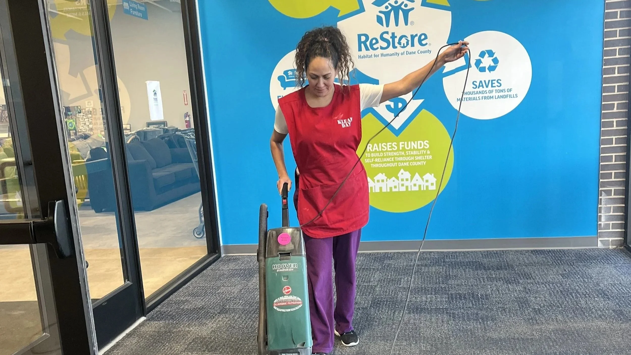 A woman in a red apron vacuuming the floor inside a building near a large blue wall with a ReStore logo and informational graphics about donations and fundraising.