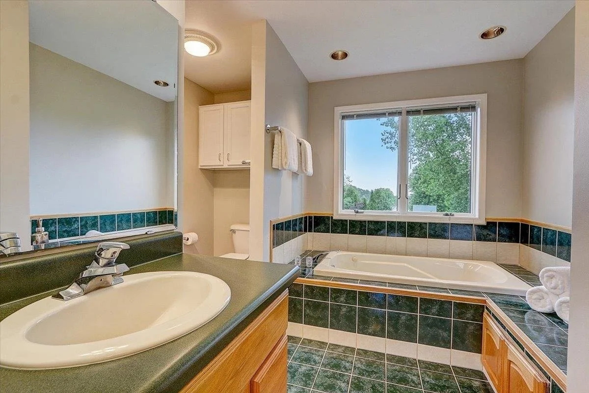 Bathroom with a sink vanity, a bathtub under a window, and a toilet, with towels hanging on a rack and green and beige tile accents.