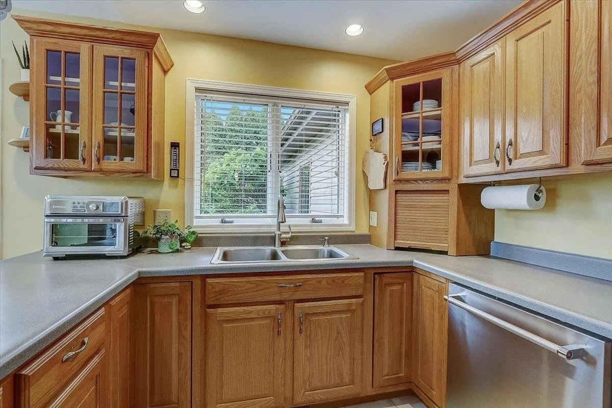 Kitchen with wooden cabinets, a double sink under a window, a countertop oven, a paper towel roll, and view of a backyard through the window.