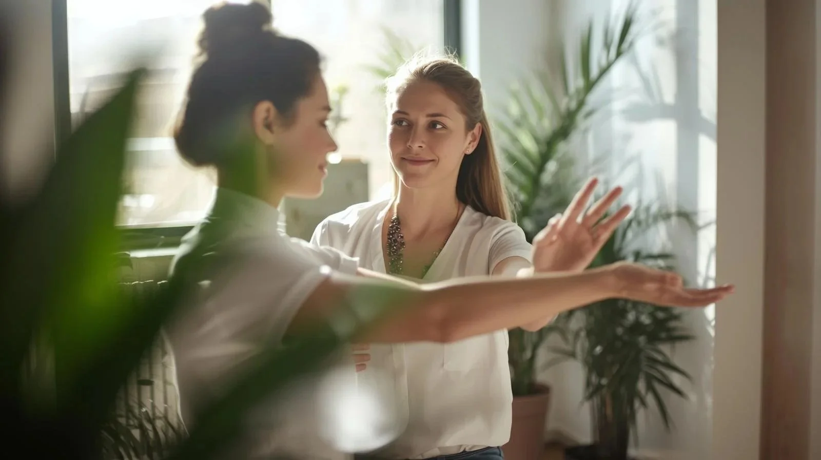 Two women are practicing a dance move or yoga pose indoors, with one woman standing and the other woman reaching out with her arms extended and palms facing each other, while they look at each other peacefully.