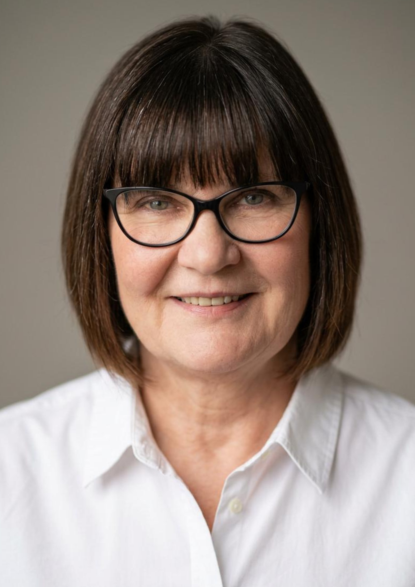 Close-up of a woman with shoulder-length brown hair wearing glasses and a white collared shirt, smiling against a plain background.