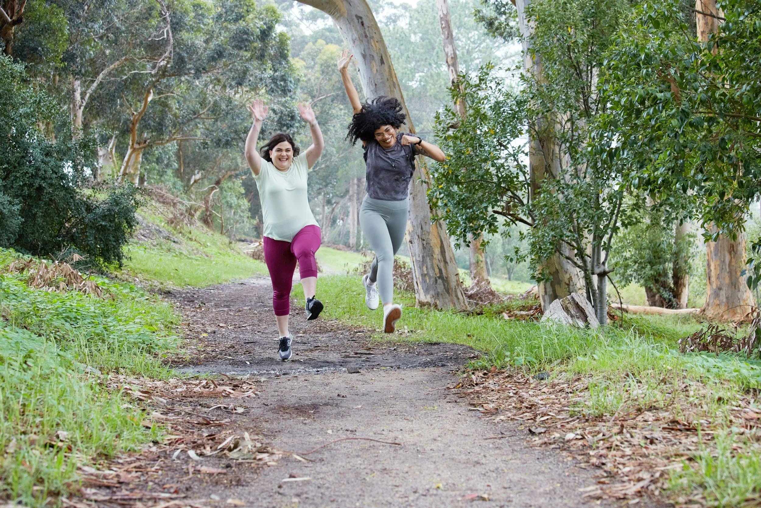 Two women joyfully running and jumping on a dirt trail in a lush, green forest.