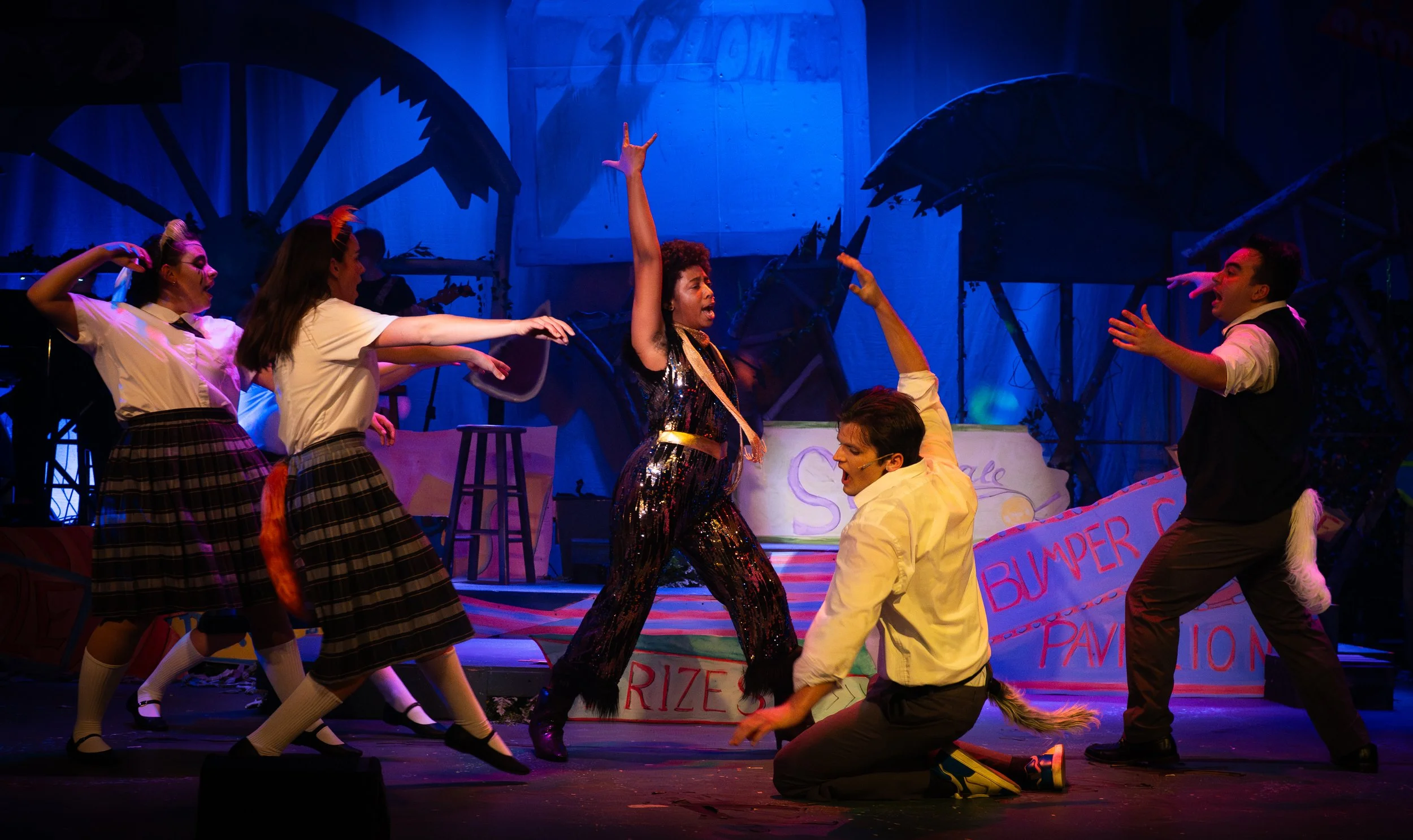 Dramatic stage scene with actors in costumes, some cats with tails, performing in a colorful, theatrical setting with props and signs.