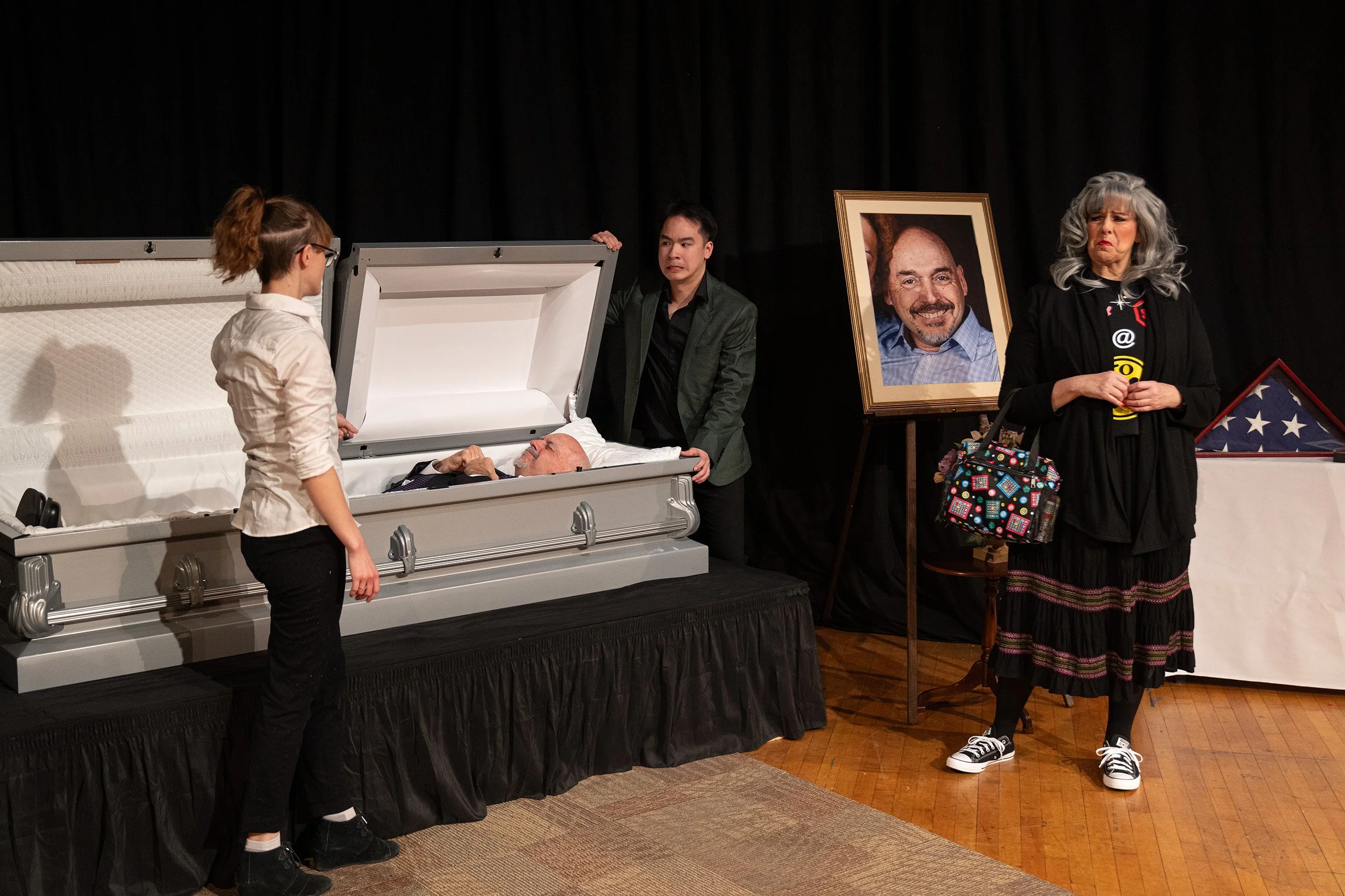 People at a memorial service, with a casket containing an elderly man, a woman with gray hair and a woman with a colorful bag standing nearby, and a large portrait of the deceased.