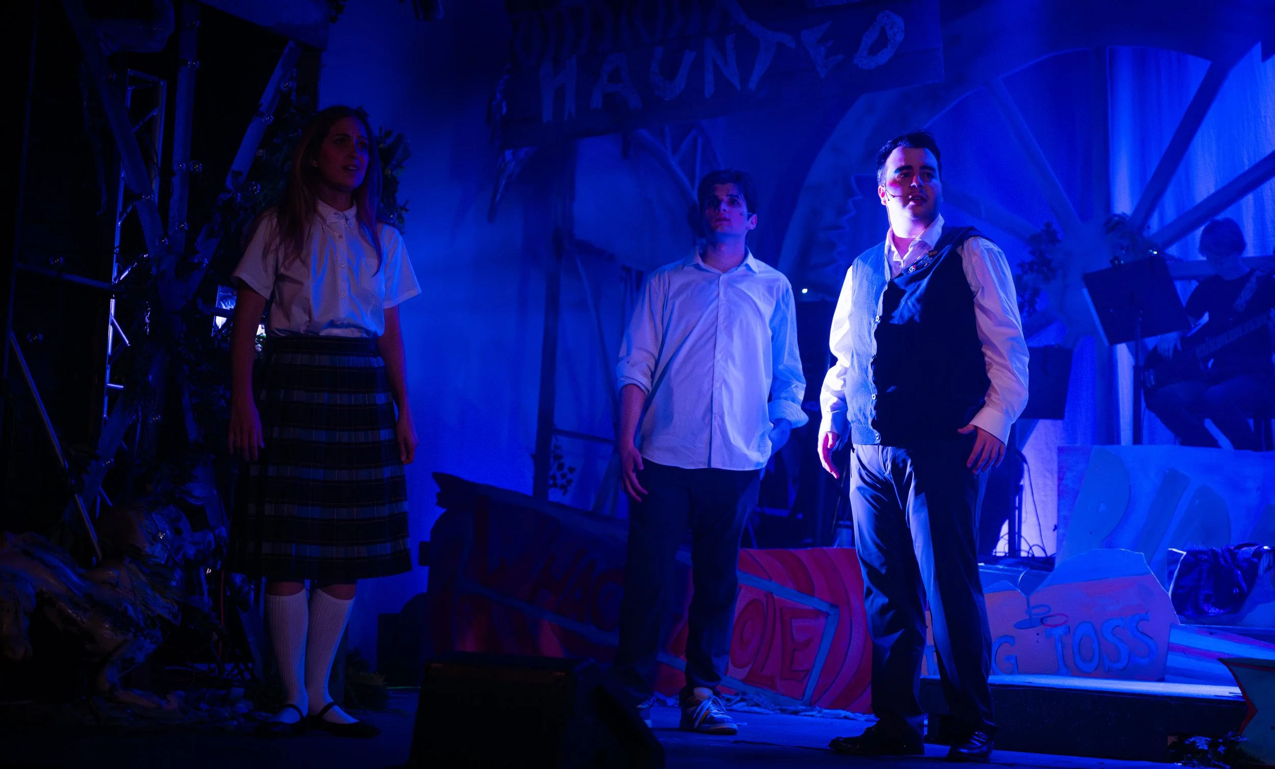 Three young actors on stage during a play, illuminated by blue lighting, with a festive Halloween-themed set background including a sign that reads 'Haunted'.