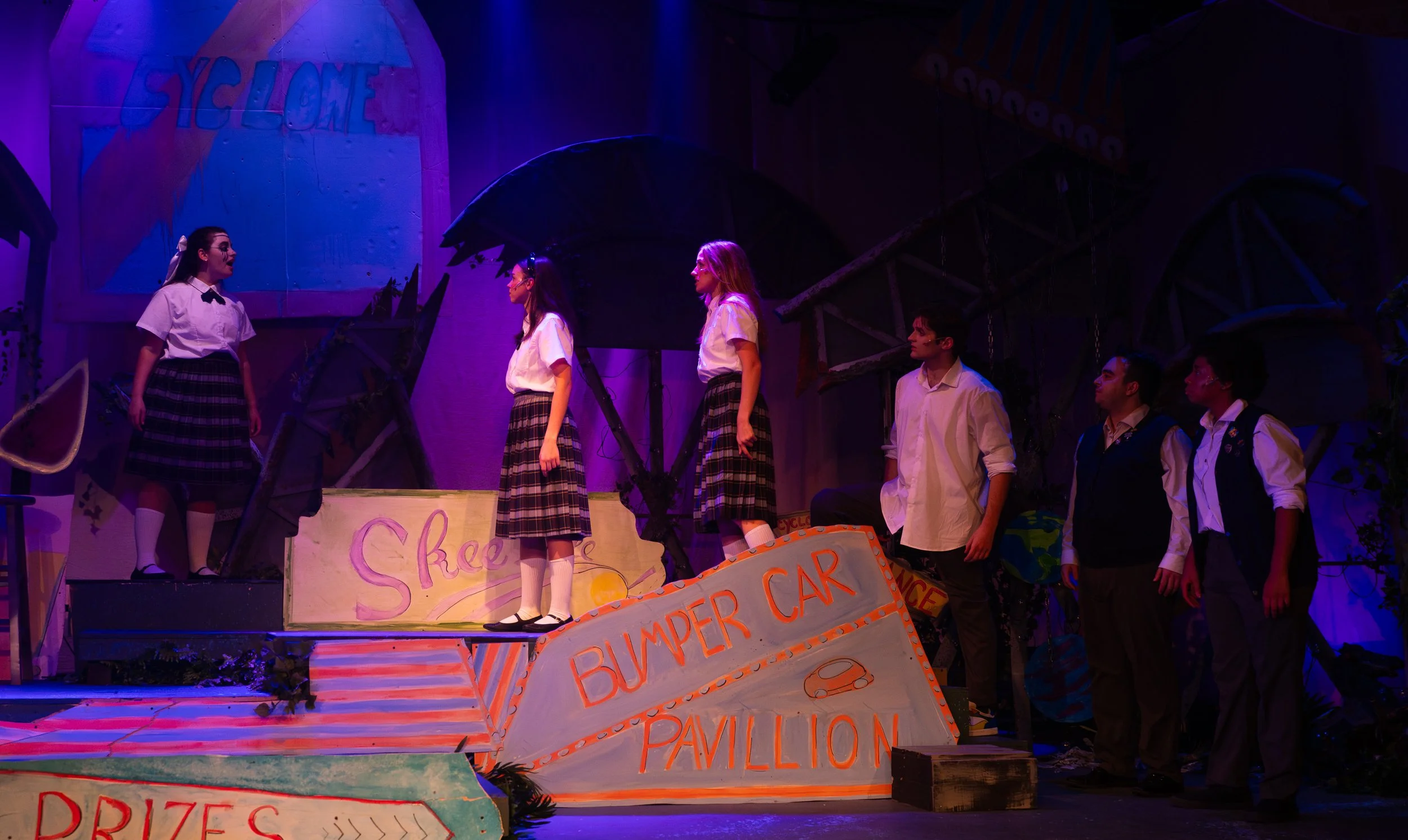 Students on stage performing in a school play, with colorful signs and set pieces resembling a carnival or amusement park, including a bumper car pavilion and prizes sign.