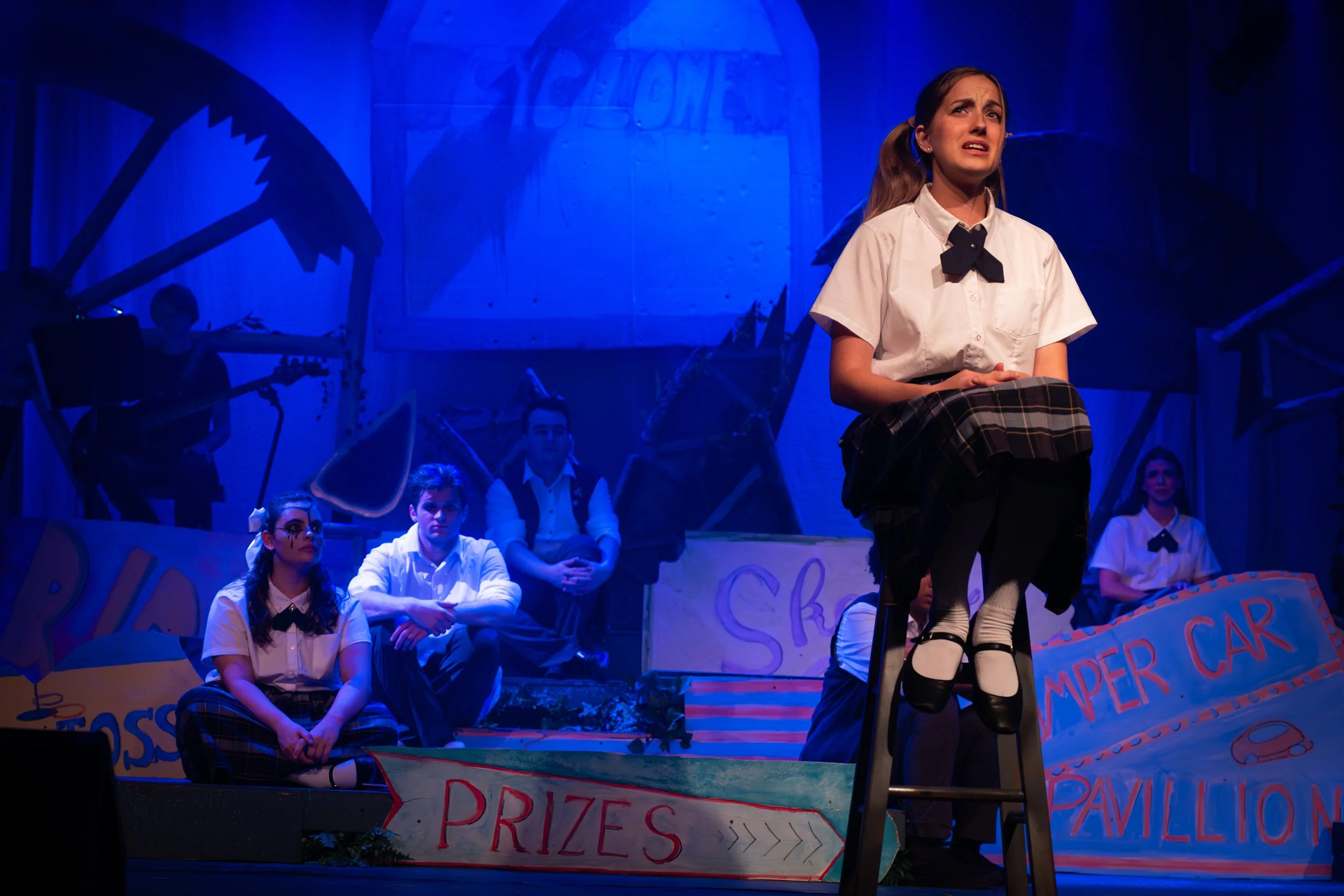 Theater scene with young actors dressed as school students. One girl standing on a chair in the foreground, looking distressed. Other students sitting and standing in the background, part of a school carnival or event setup with signs for prizes, a b