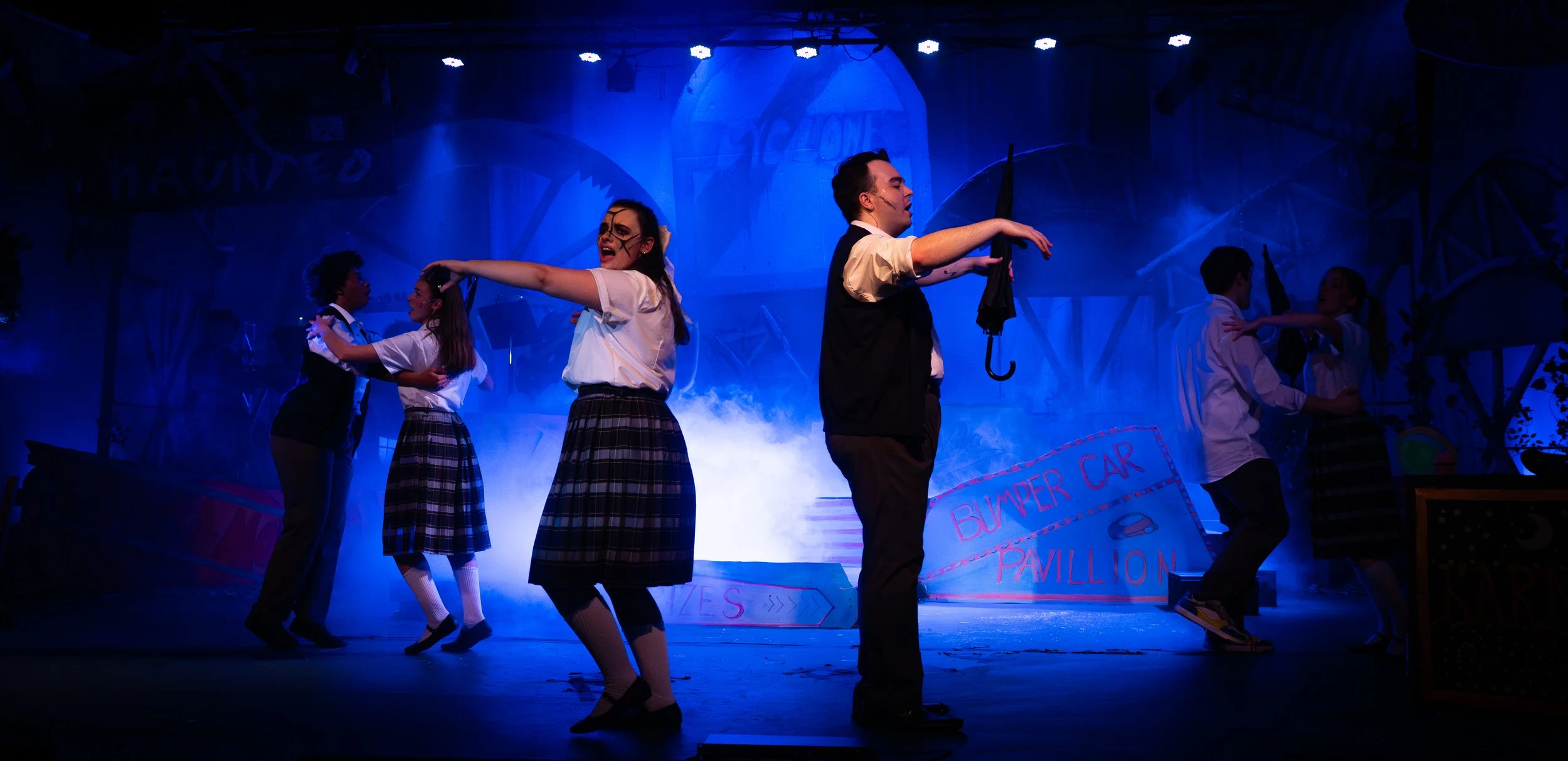 Theatrical stage scene with young performers in school uniforms, performing a dance with props like umbrellas, under blue lighting with fog effects and amusement park or carnival set decorations.