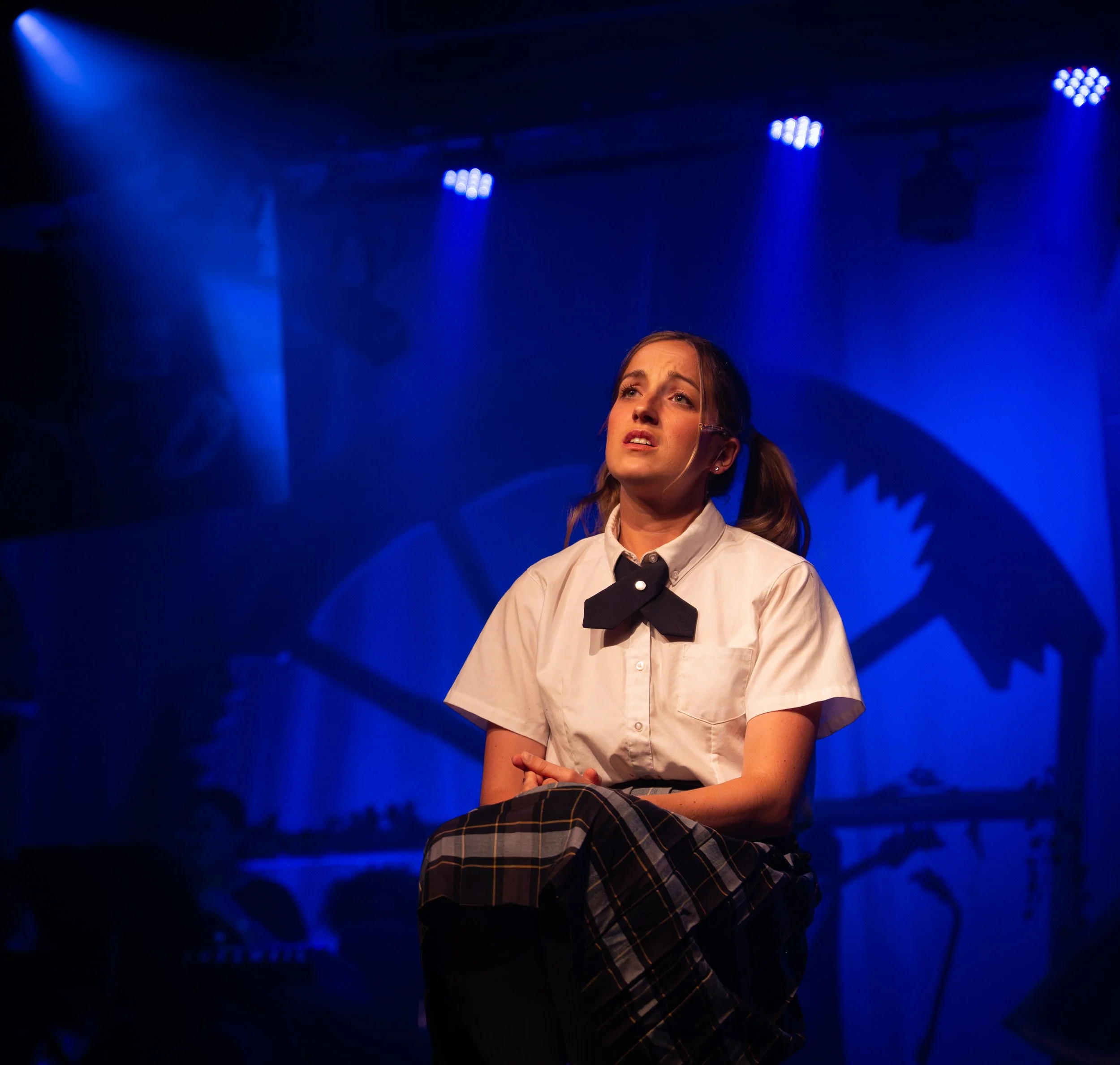 A young woman in a school uniform performing on stage, looking emotional, with blue stage lighting and abstract background art.