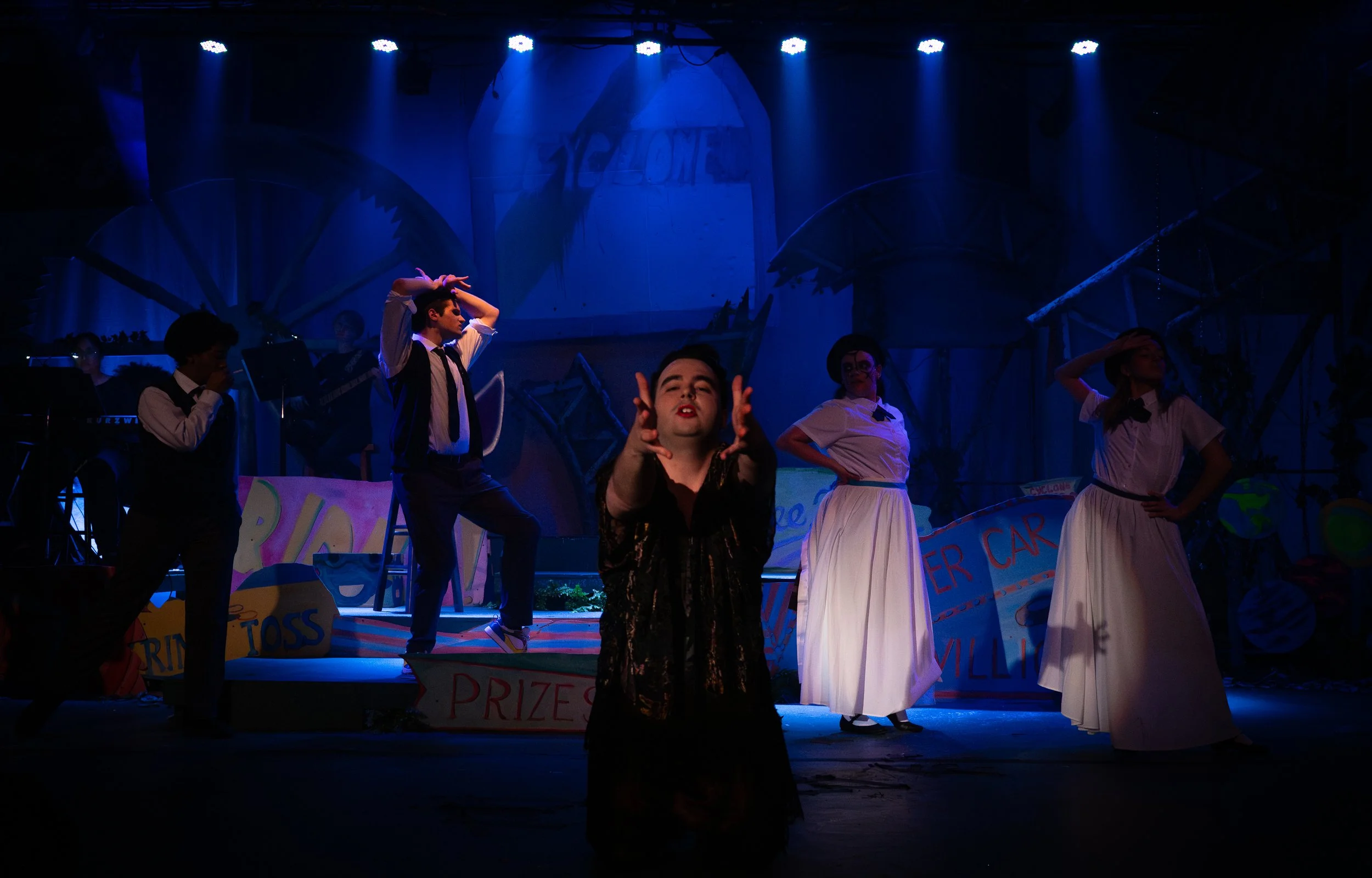 People performing on stage with colorful signs and decorations, including a sign that says 'PRIZES' and another that says 'ROLLER CAR,' under blue stage lighting.