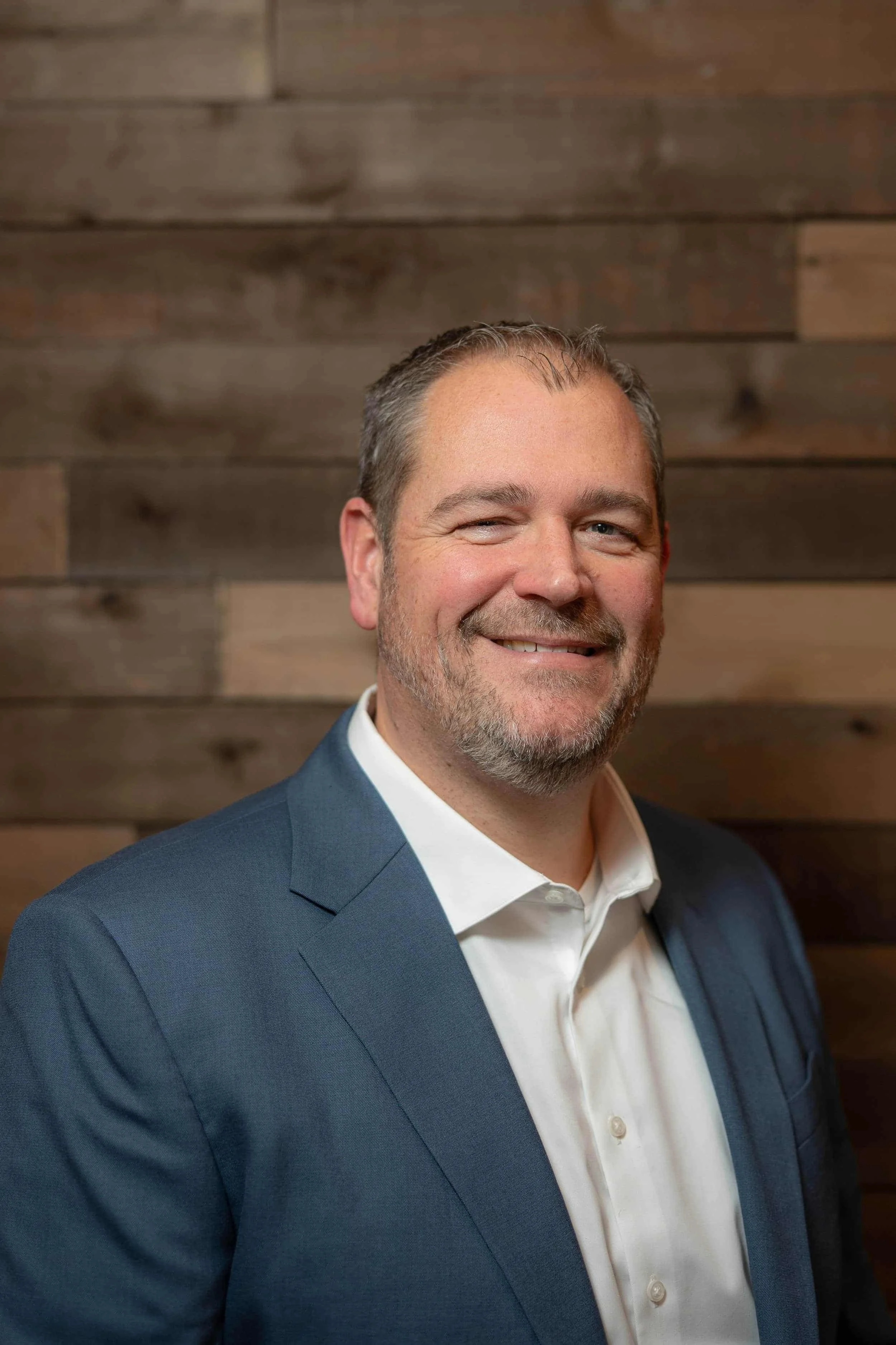 Headshot of a middle-aged man with short hair, a beard, and a mustache, smiling in a blue suit and white shirt, standing against a wooden background.