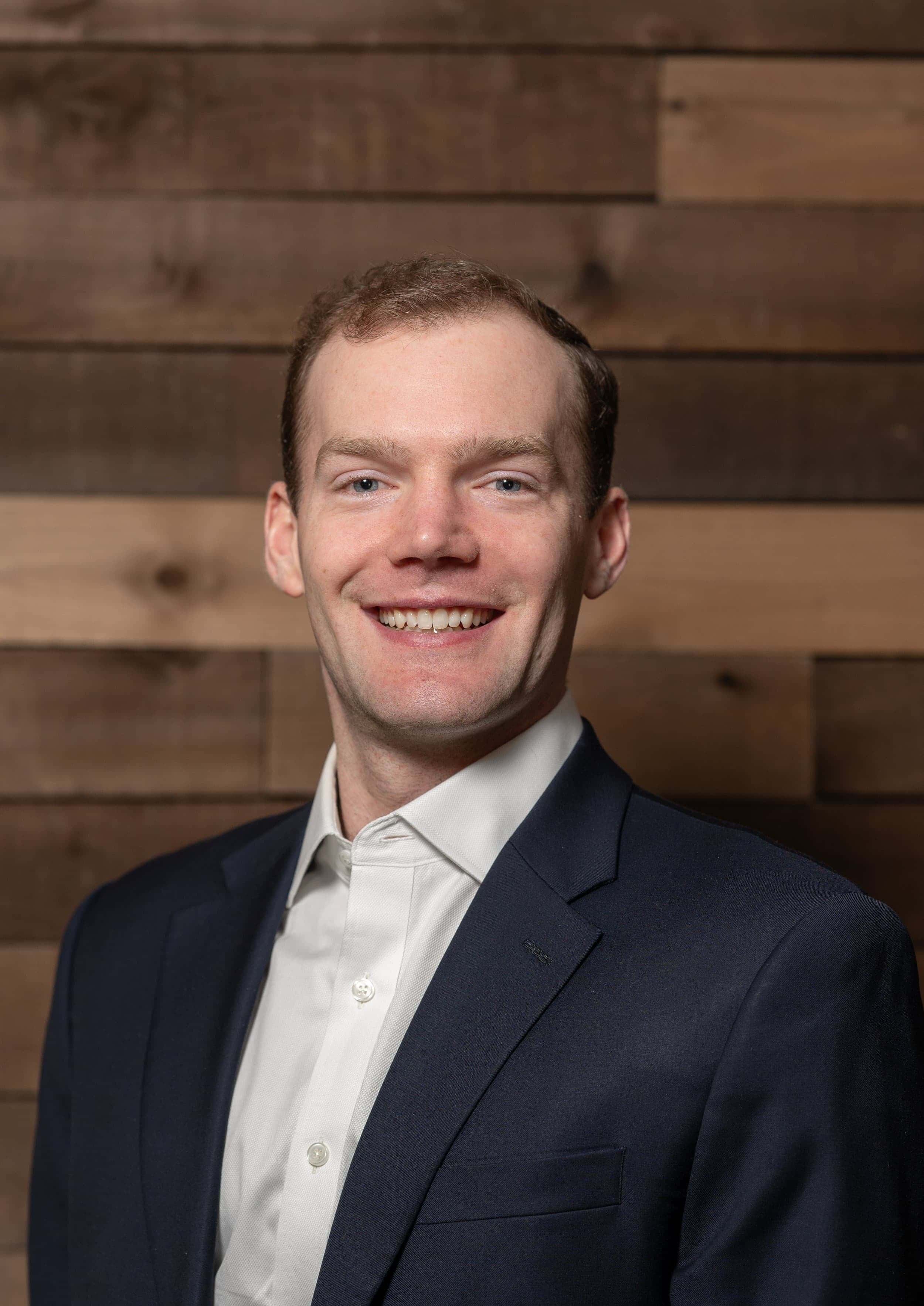 Portrait of a young man in a white shirt and navy blazer smiling in front of a wooden wall background.