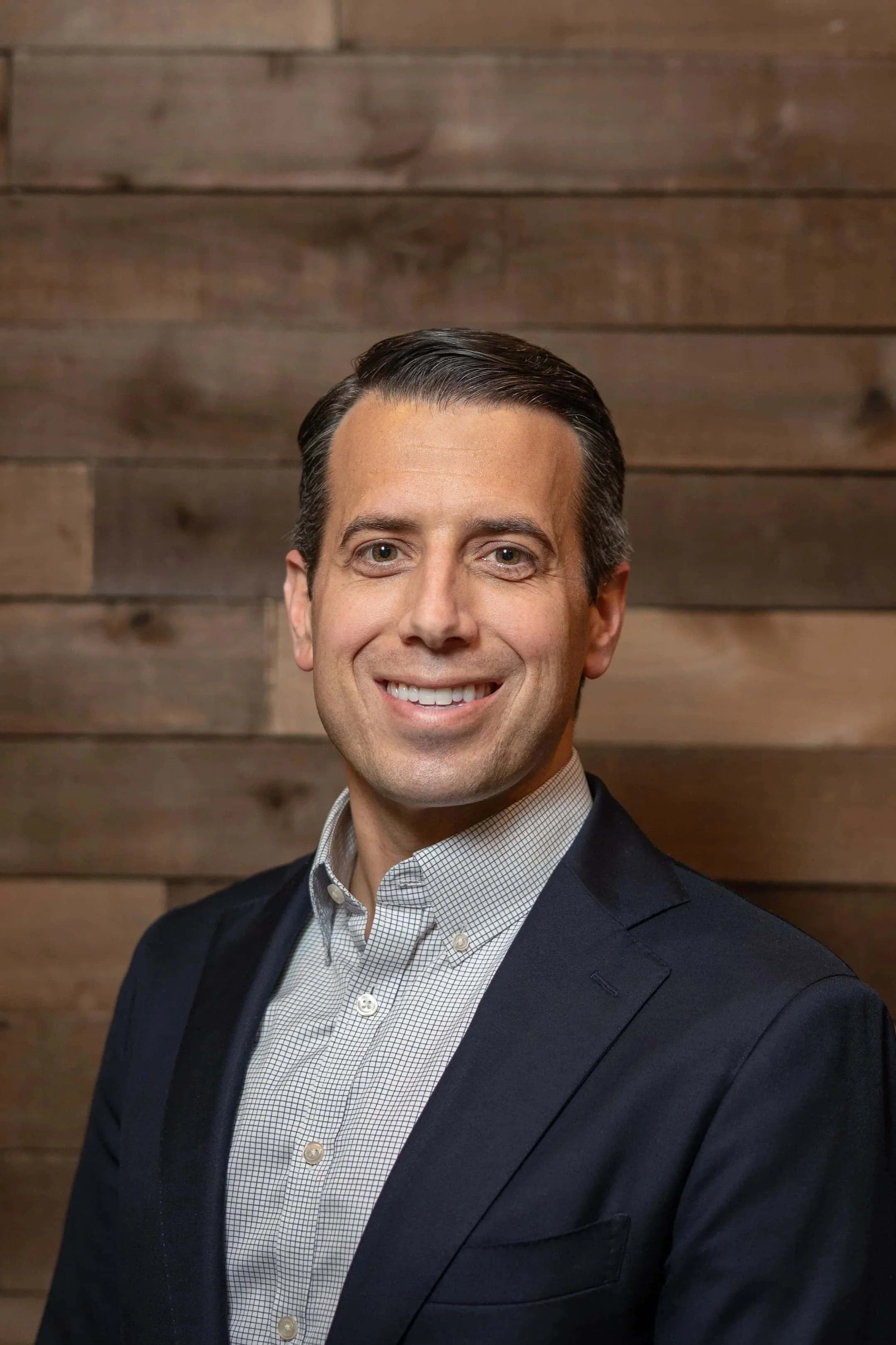 Portrait of a smiling man with dark hair, wearing a dark blazer and a light-colored button-up shirt, standing in front of a wooden wall.