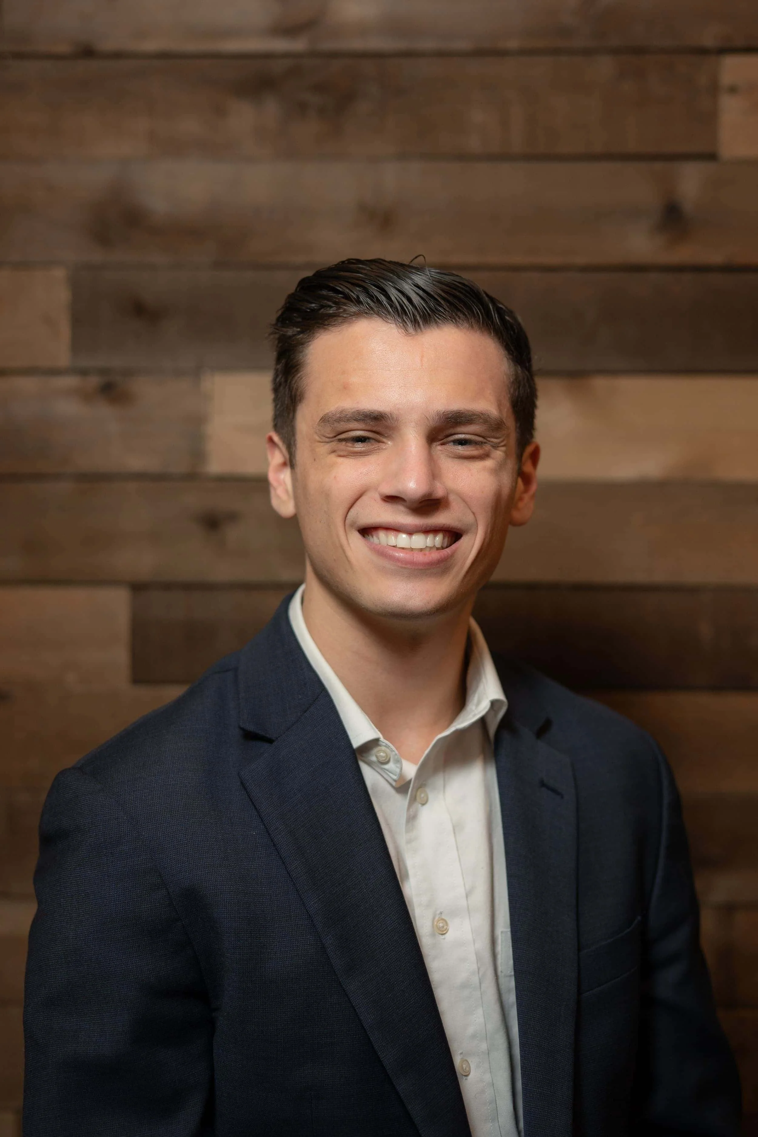 A young man with dark hair, wearing a white shirt and a dark blazer, smiling in front of a wooden wall.