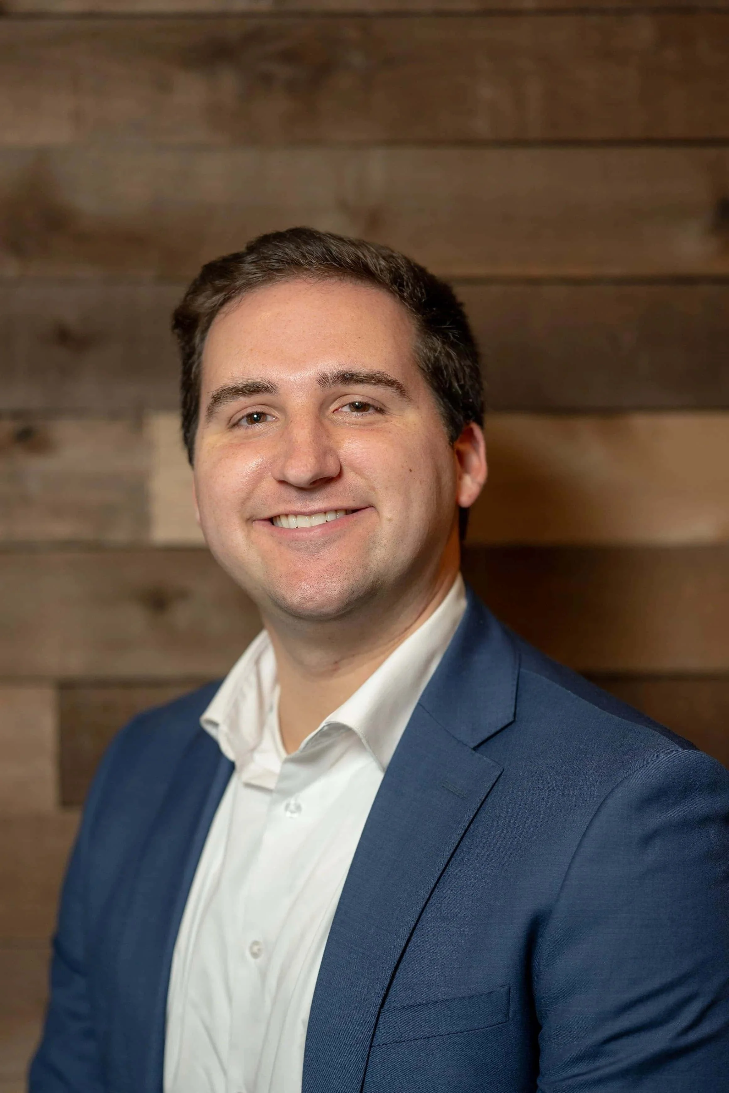 A smiling man in a navy blazer and white shirt standing in front of a wooden background.