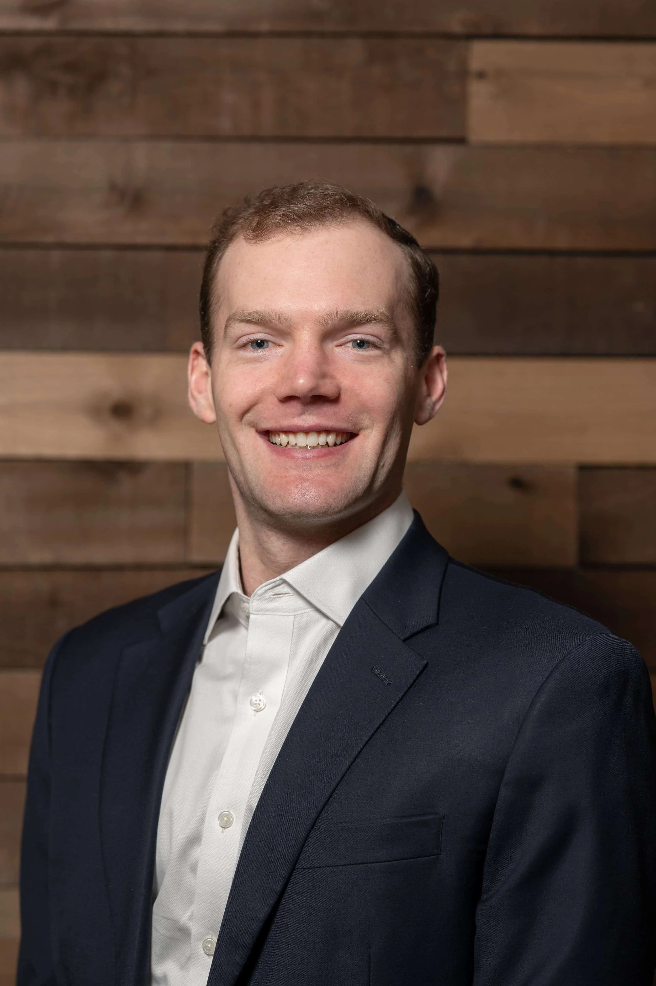 Headshot of a smiling man with short brown hair wearing a navy suit and white shirt, standing against a wooden wall.